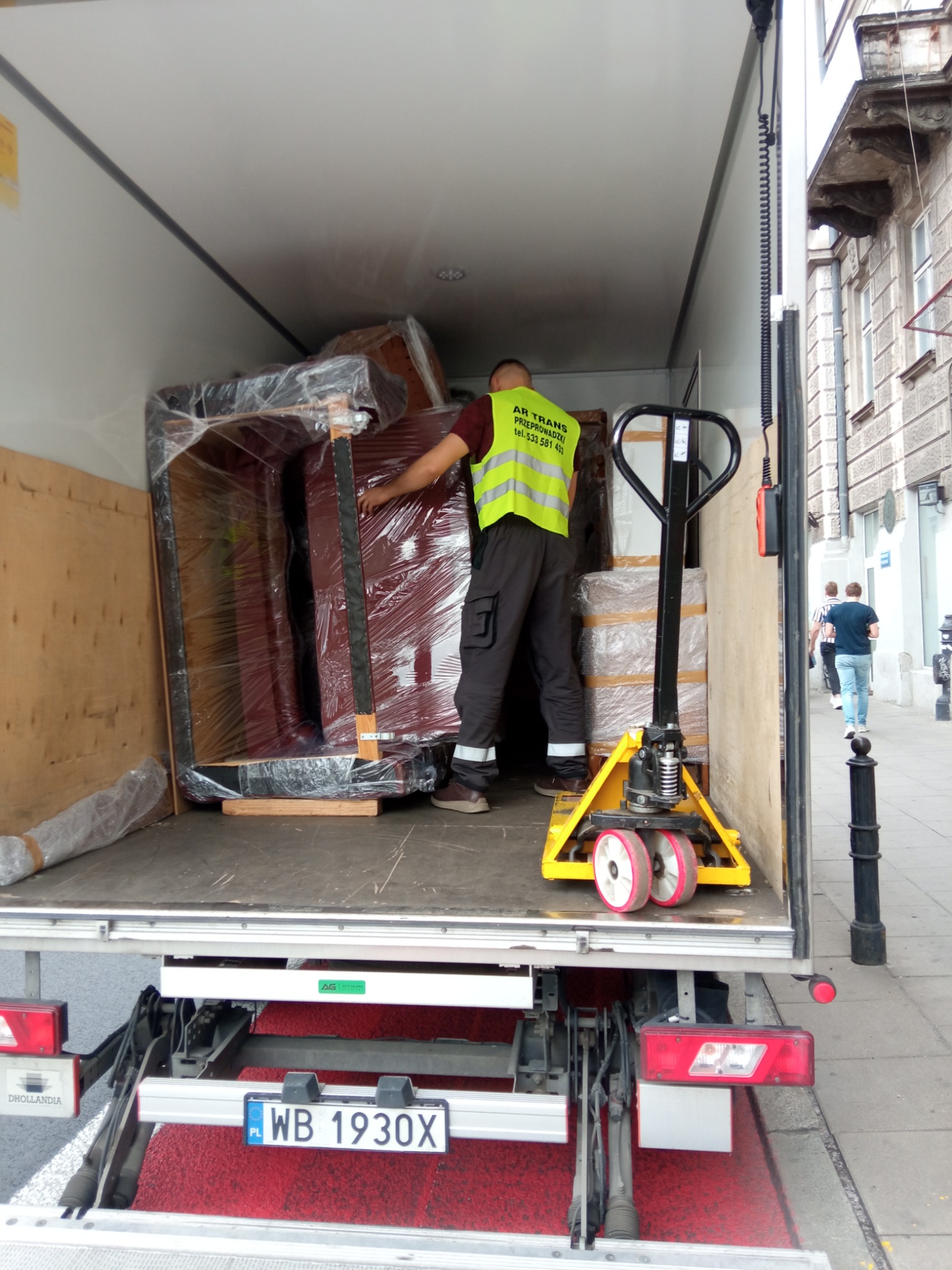 Pracownik w kamizelce AR TRANS zabezpiecza meble owinięte folią stretch wewnątrz furgonetki z podnośnikiem, widoczny wózek paletowy i tablica rejestracyjna WB 1930X.
