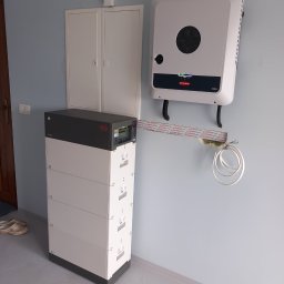 SkyVolt Rafał Kornas - Biały magazyn energii Byd Premium LVL 15.4 i falownik Fronius Symo zamontowane na ścianie wewnątrz budynku, widoczne przewody i rurki instalacyjne.