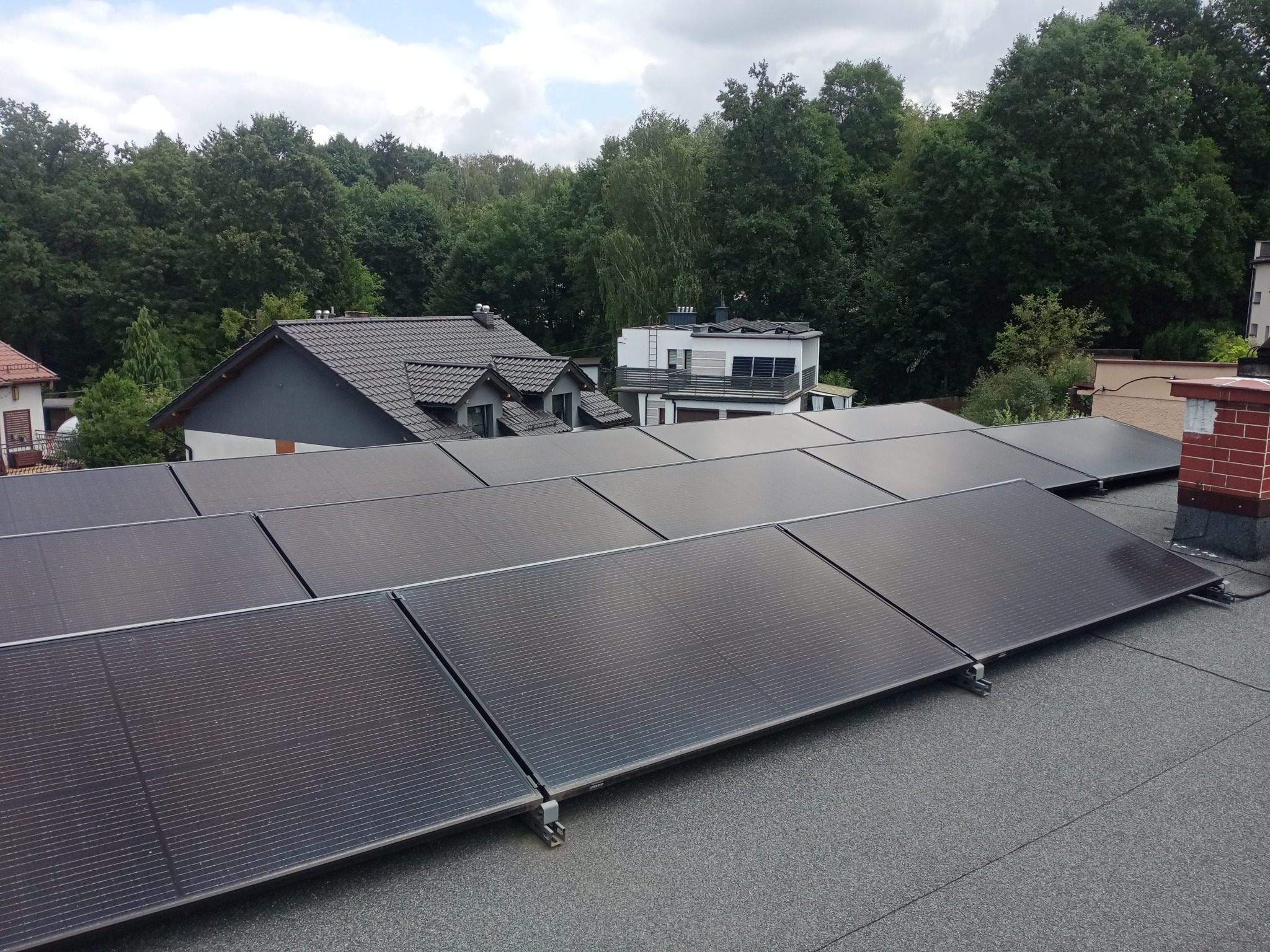 Rząd paneli słonecznych na płaskim dachu domu jednorodzinnego, widok z góry, w tle drzewa i budynki.