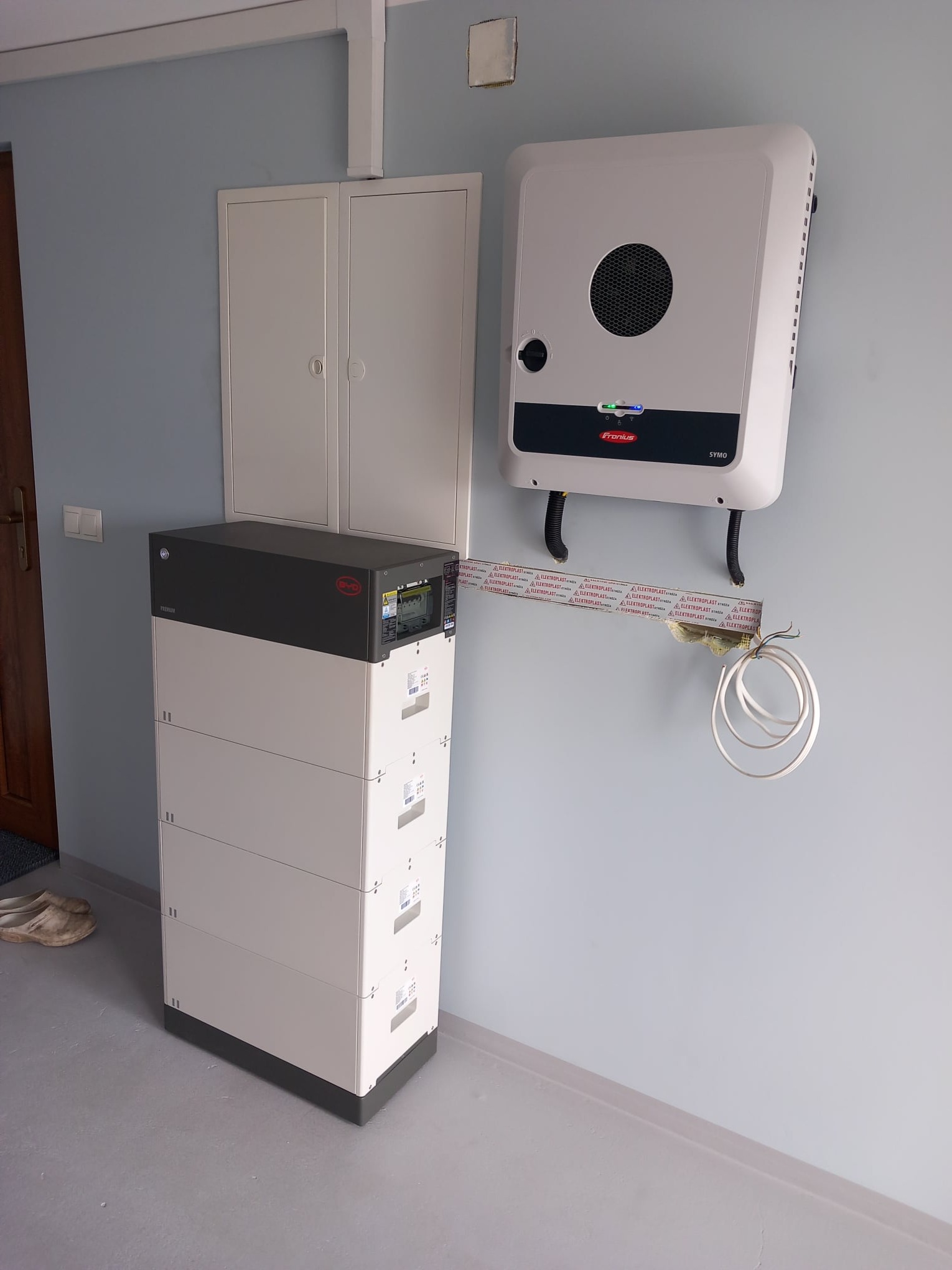 Biały magazyn energii Byd Premium LVL 15.4 i falownik Fronius Symo zamontowane na ścianie wewnątrz budynku, widoczne przewody i rurki instalacyjne.