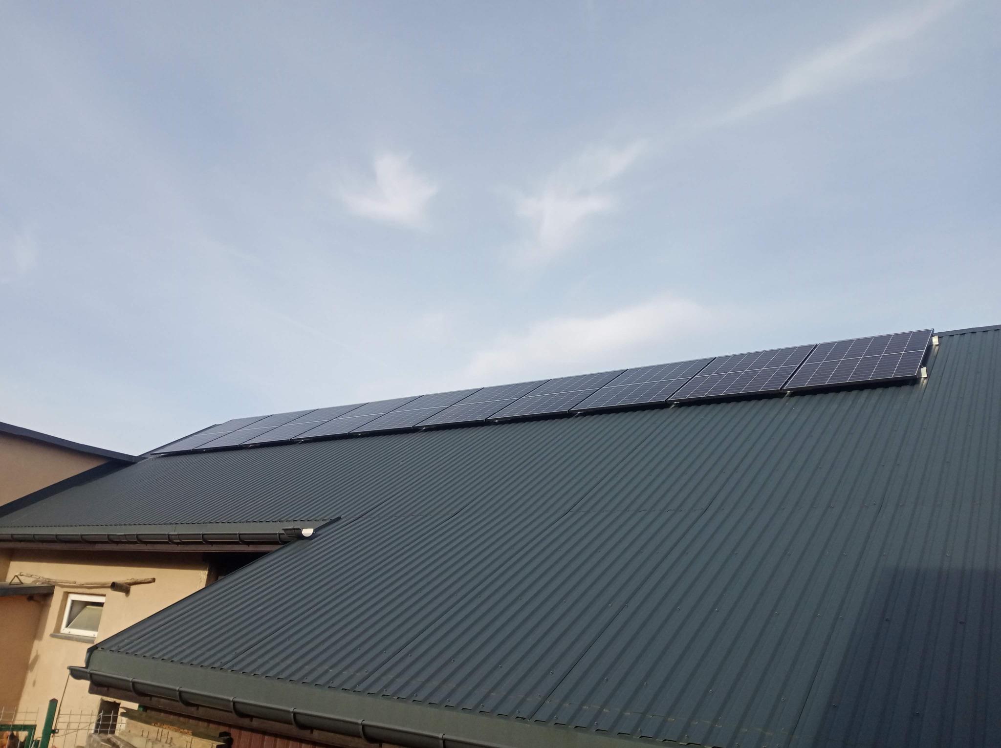 Rząd paneli słonecznych na ciemnoszarym dachu z blachy trapezowej; widok z dołu, błękitne niebo z delikatnymi obłokami w tle.