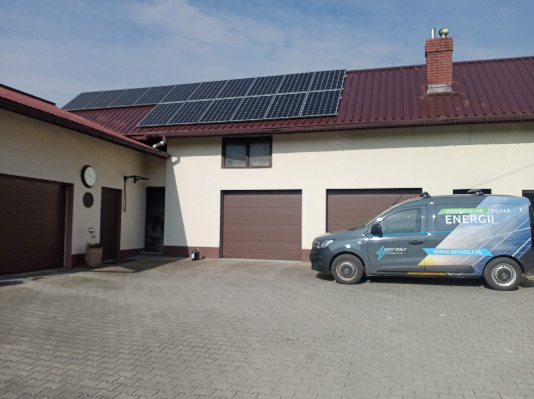 Dom jednorodzinny z panelami słonecznymi na dachu i samochodem firmowym z logo na podjeździe z kostki brukowej.