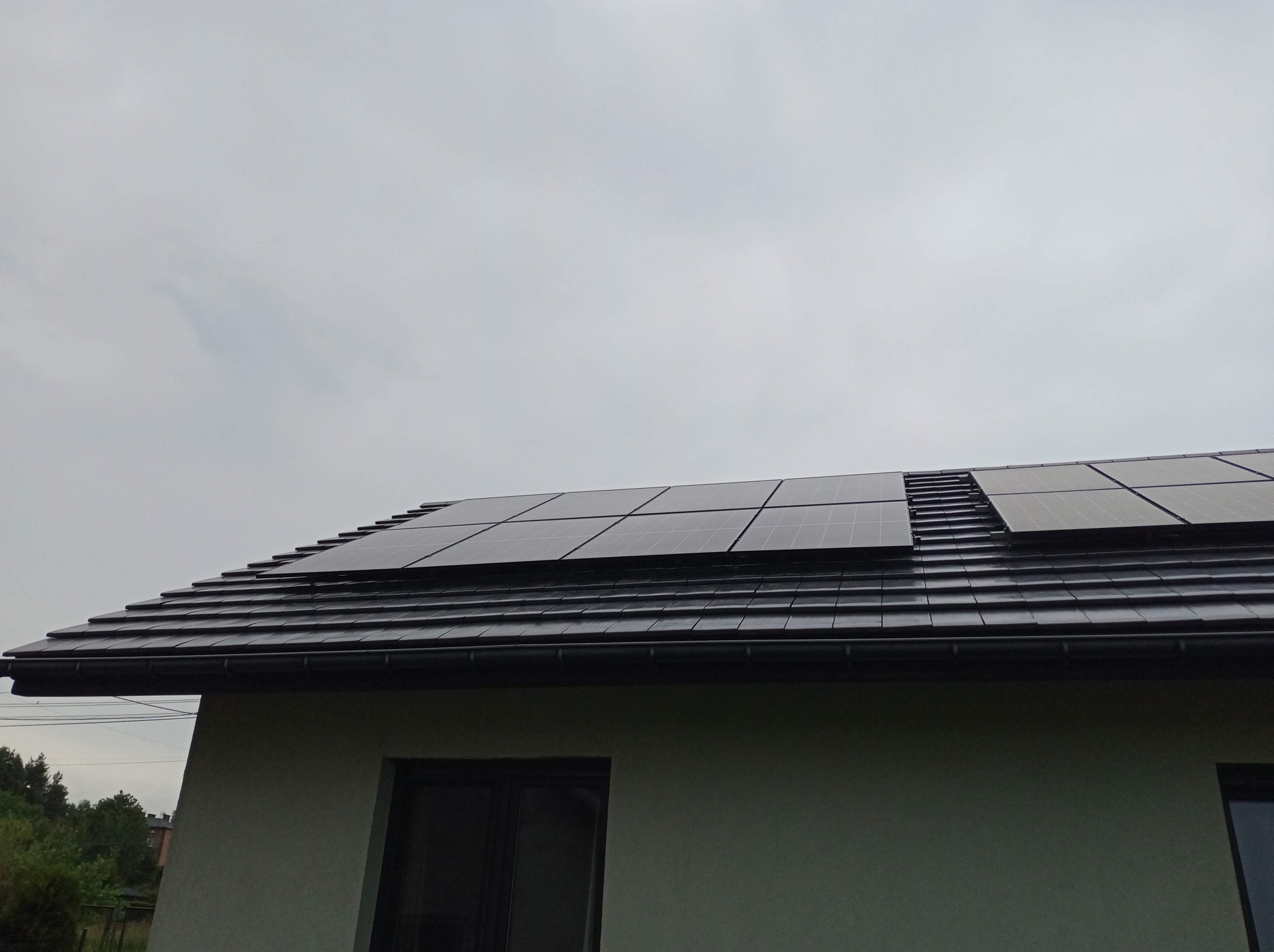 Panele słoneczne zamontowane na dachu domu pokrytego ciemną dachówką, widok z dołu na tle pochmurnego nieba.