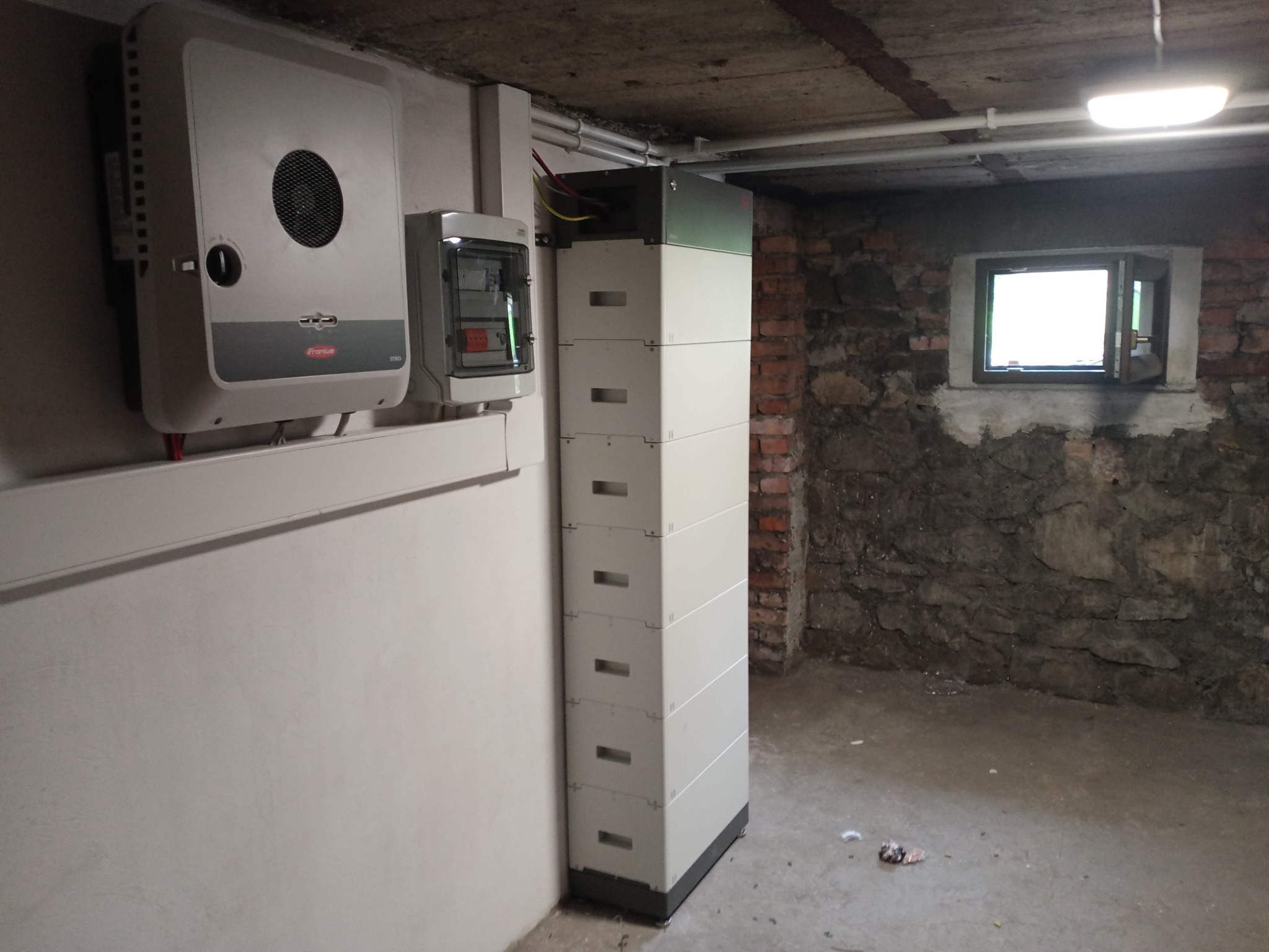 Instalacja magazynu energii składającego się z kilku modułów w piwnicy z kamiennymi ścianami, obok falownik Fronius i skrzynka elektryczna.