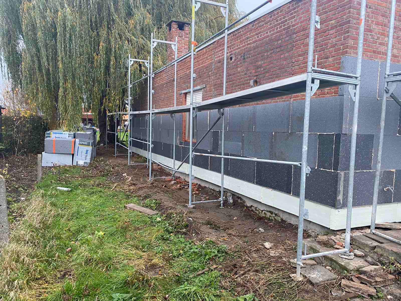 Ocieplanie elewacji domu z cegły, widoczne rusztowanie, styropian grafitowy i zapas materiału pod wierzbą płaczącą.