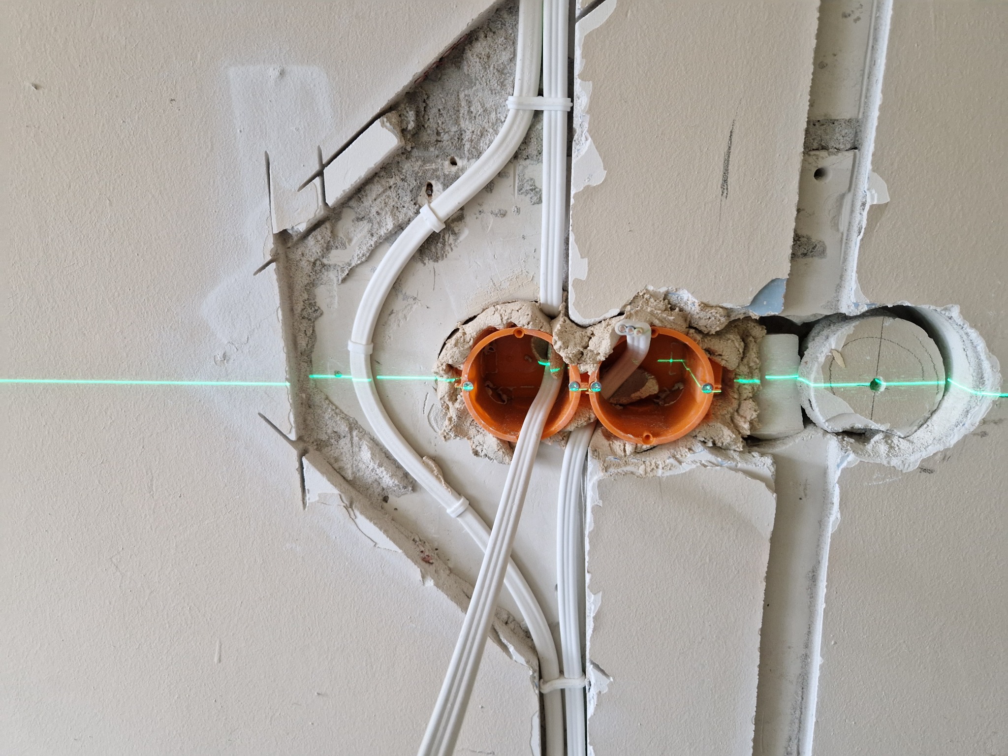Dwa pomarańczowe puszki podtynkowe osadzone w ścianie, z widocznymi przewodami elektrycznymi i poziomą linią lasera wskazującą poziom. Ściana w stanie surowym, z widocznymi śladami po kuciu i gładzią.