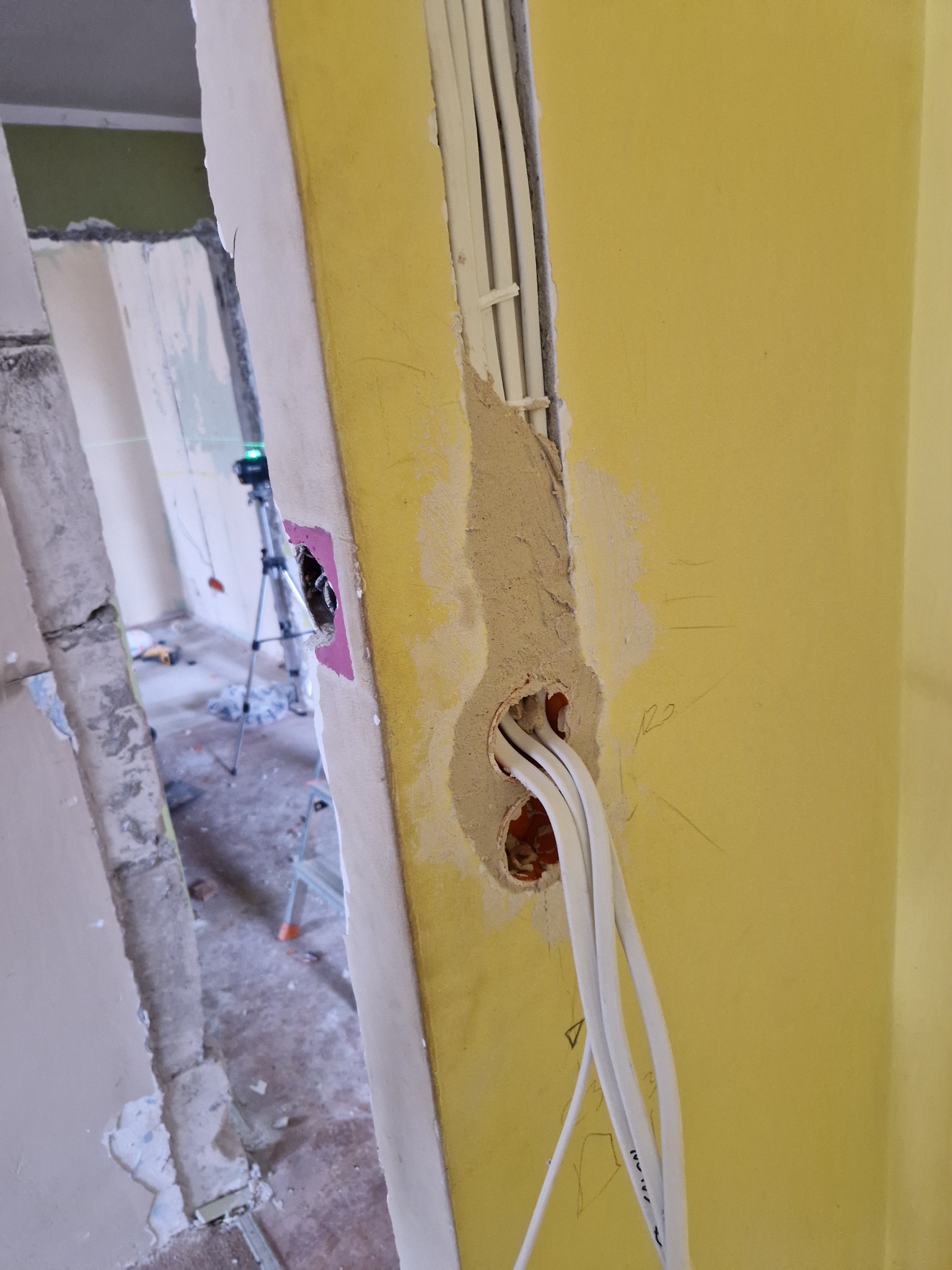 Żółta ściana z wywierconymi otworami i wystającymi przewodami elektrycznymi, ślady po wierceniu i szpachlowaniu, w tle widoczny laser krzyżowy na statywie w remontowanym pomieszczeniu.