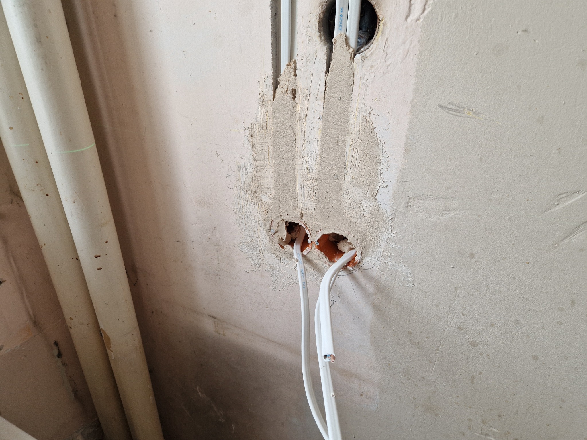 Ściana w trakcie remontu z widocznymi przewodami elektrycznymi wystającymi z wywierconych otworów, świeżo nałożona zaprawa gipsowa w miejscach bruzd po przewodach.