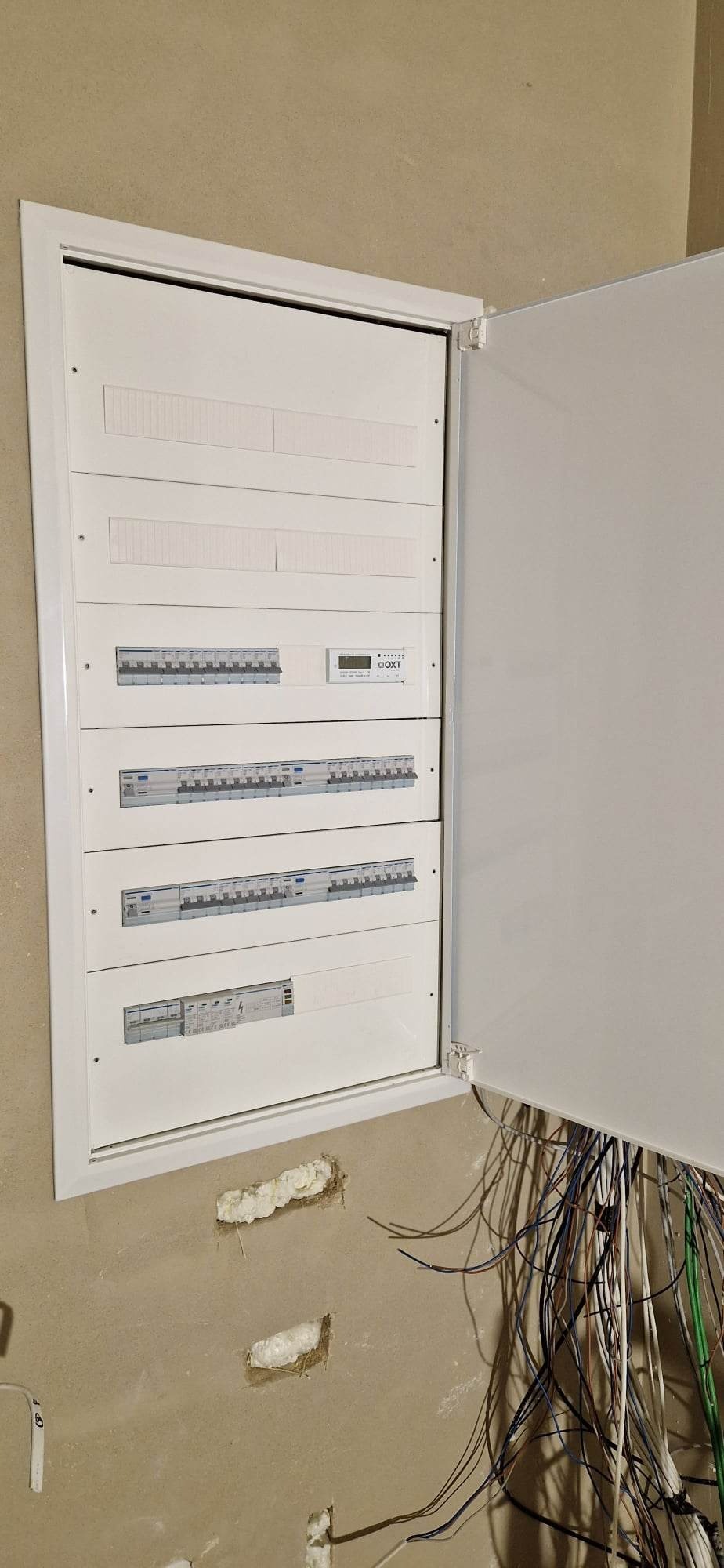 Otwarta szafka elektryczna z widocznymi bezpiecznikami i przewodami wychodzącymi ze ściany, w trakcie montażu.