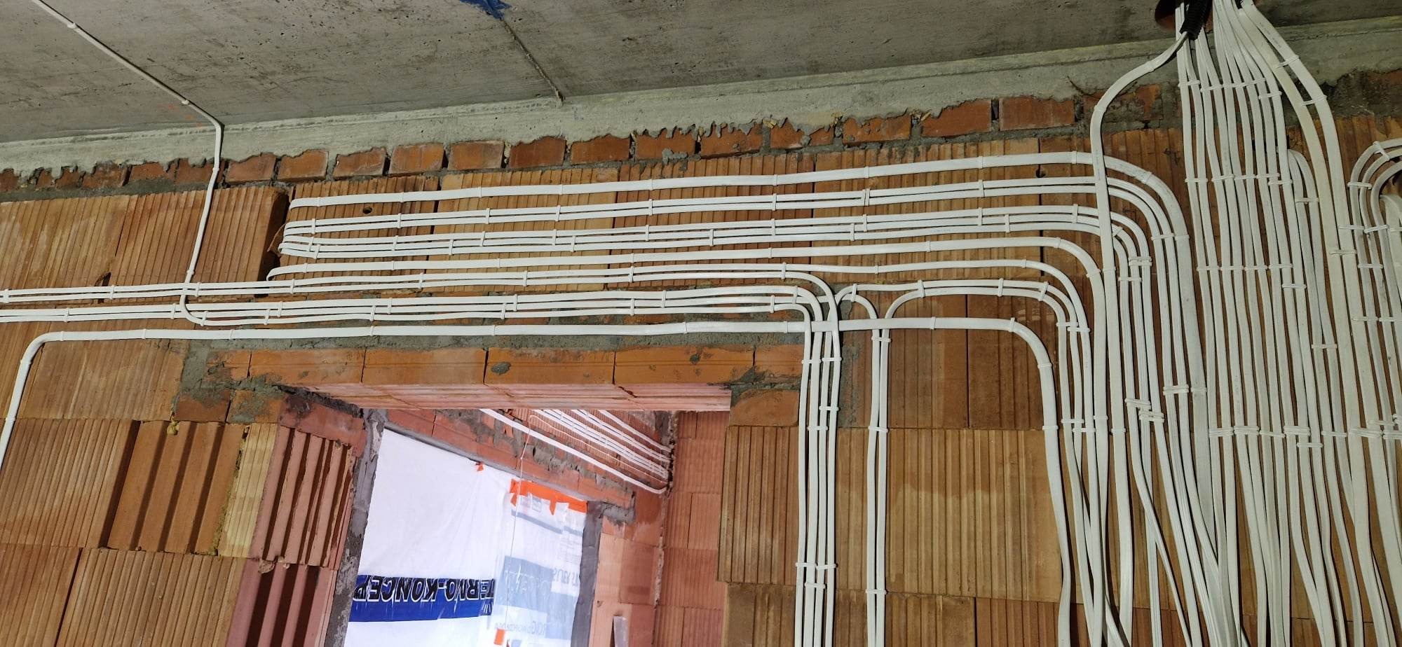 Białe peszle elektryczne ułożone równolegle, przymocowane do surowej ściany z czerwonej cegły w trakcie instalacji, widoczne przejście przez otwór drzwiowy.