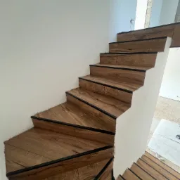 Wykończone drewnopodobne schody z czarnymi krawędziami na białym tle ściany, widoczne niedoskonałości wykończenia przy ścianie i zabezpieczona podłoga na górze schodów.