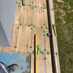 Układanie płytek imitujących drewno na balkonie, widoczne narzędzia glazurnicze, poziomice, krzyżyki dystansowe, klej i fragment butów glazurnika w rogu kadru.