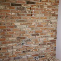 Ściana wyłożona dekoracyjną cegłą w odcieniach czerwieni i brązu, z widocznymi przewodami elektrycznymi wystającymi ze ściany, szara podłoga i fragment mebla w prawym dolnym rogu.