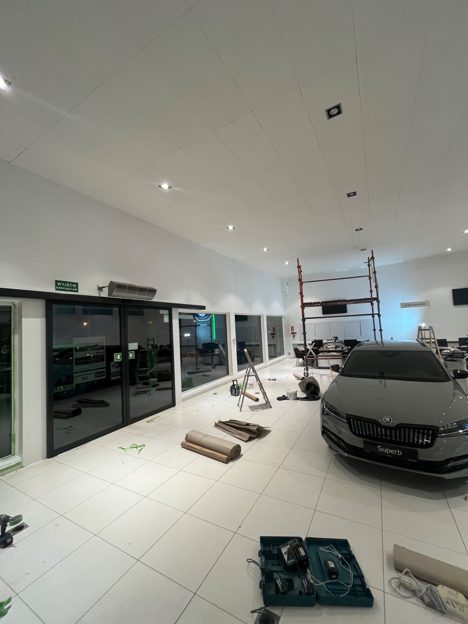 Montaż sufitu podwieszanego w salonie samochodowym, widoczne rusztowanie, płyty gipsowo-kartonowe, narzędzia oraz zaparkowany samochód Skoda Superb.