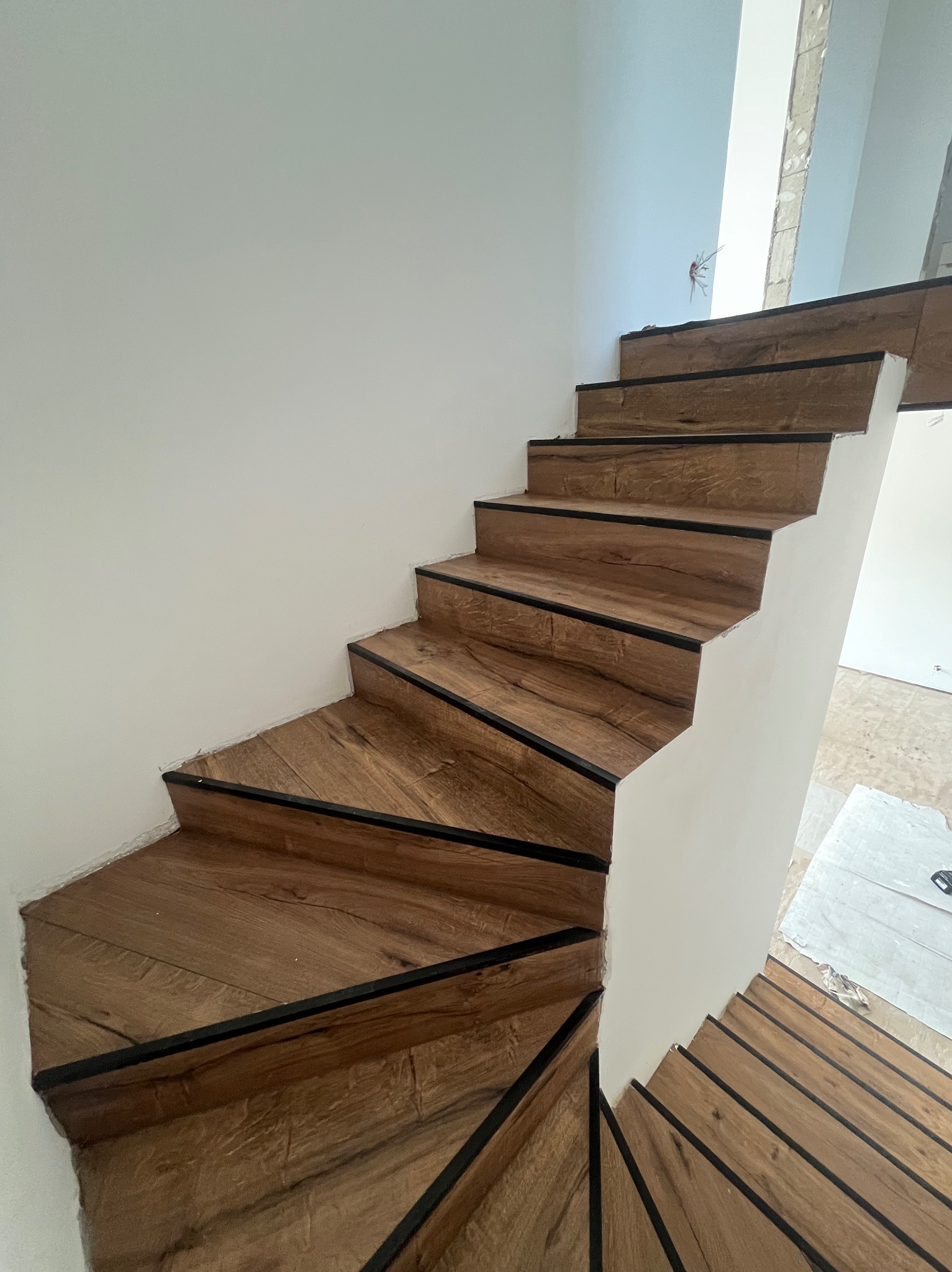 Wykończone drewnopodobne schody z czarnymi krawędziami na białym tle ściany, widoczne niedoskonałości wykończenia przy ścianie i zabezpieczona podłoga na górze schodów.
