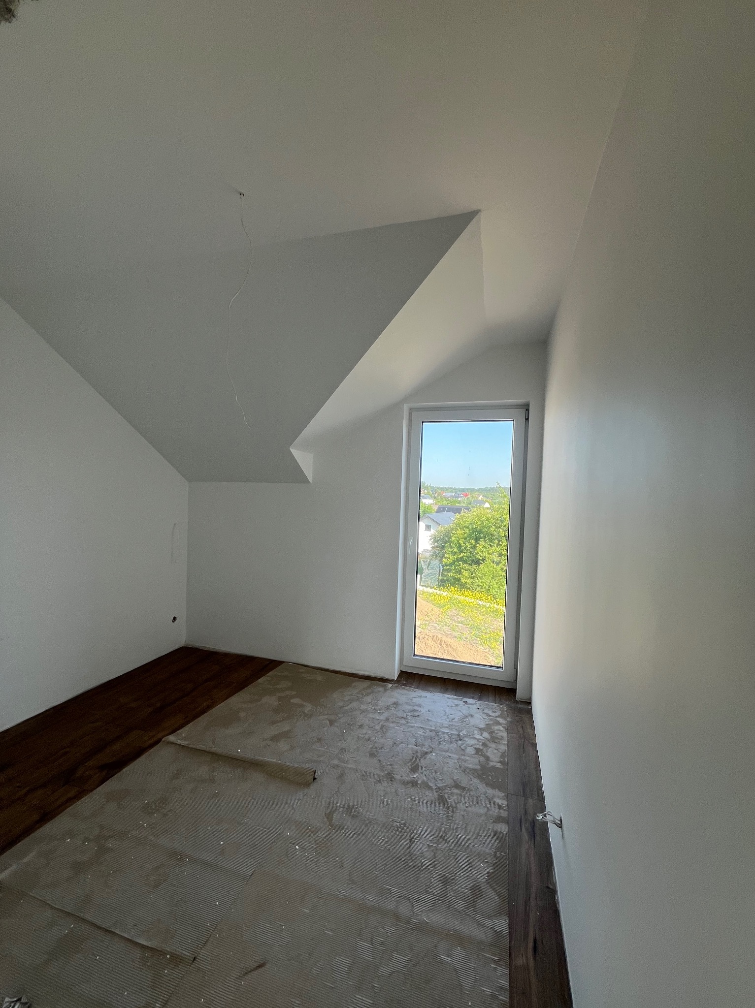 Pokój na poddaszu w trakcie układania paneli podłogowych; widoczne fragmenty ułożonej podłogi oraz podkład pod panele, świeżo pomalowane ściany na biało, okno z widokiem na zieleń.