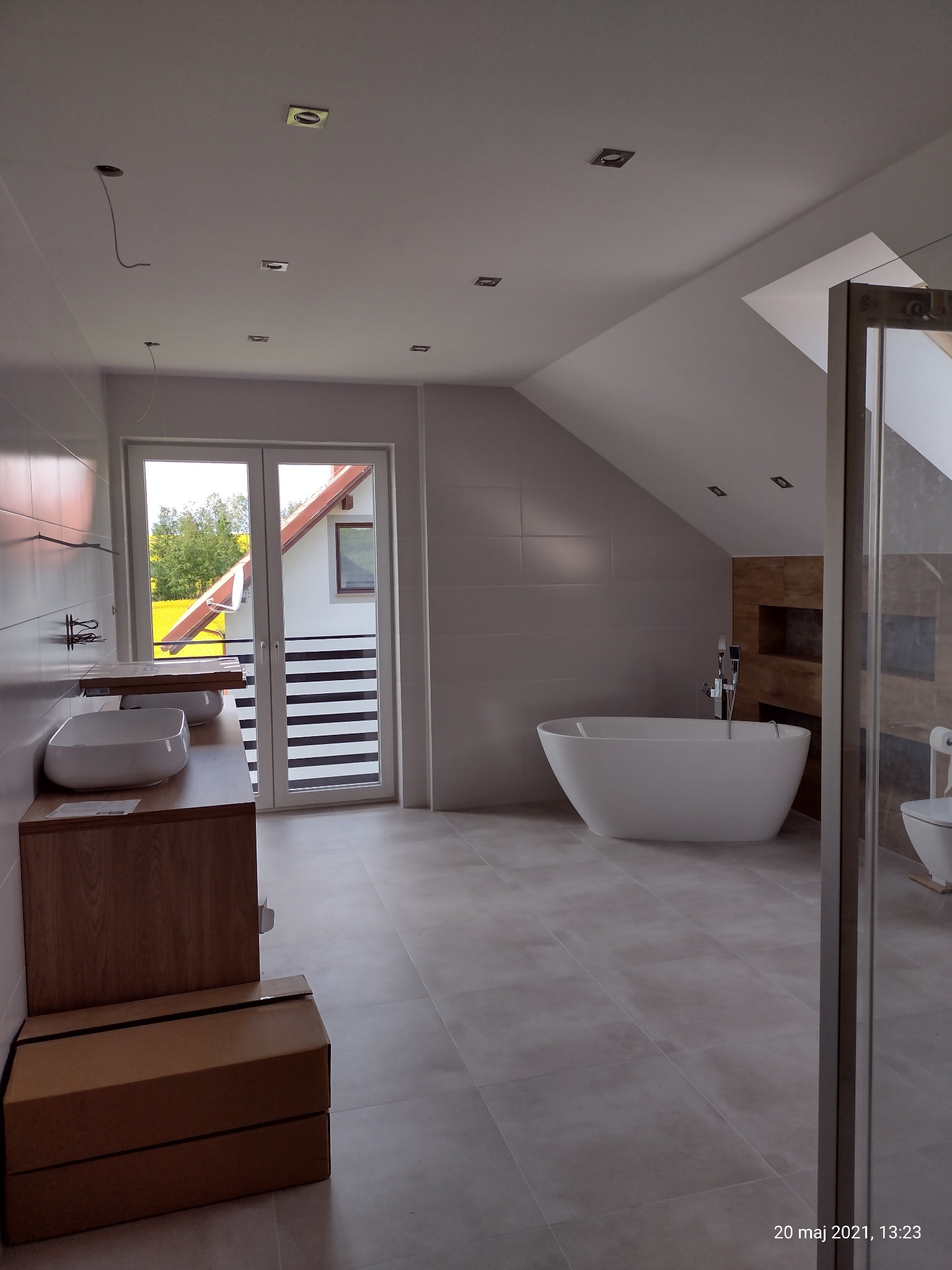 Przestronna łazienka na poddaszu z wolnostojącą wanną, dwiema umywalkami na drewnianym blacie, kabiną prysznicową ze szklanymi drzwiami i oknem balkonowym z widokiem na zieleń.