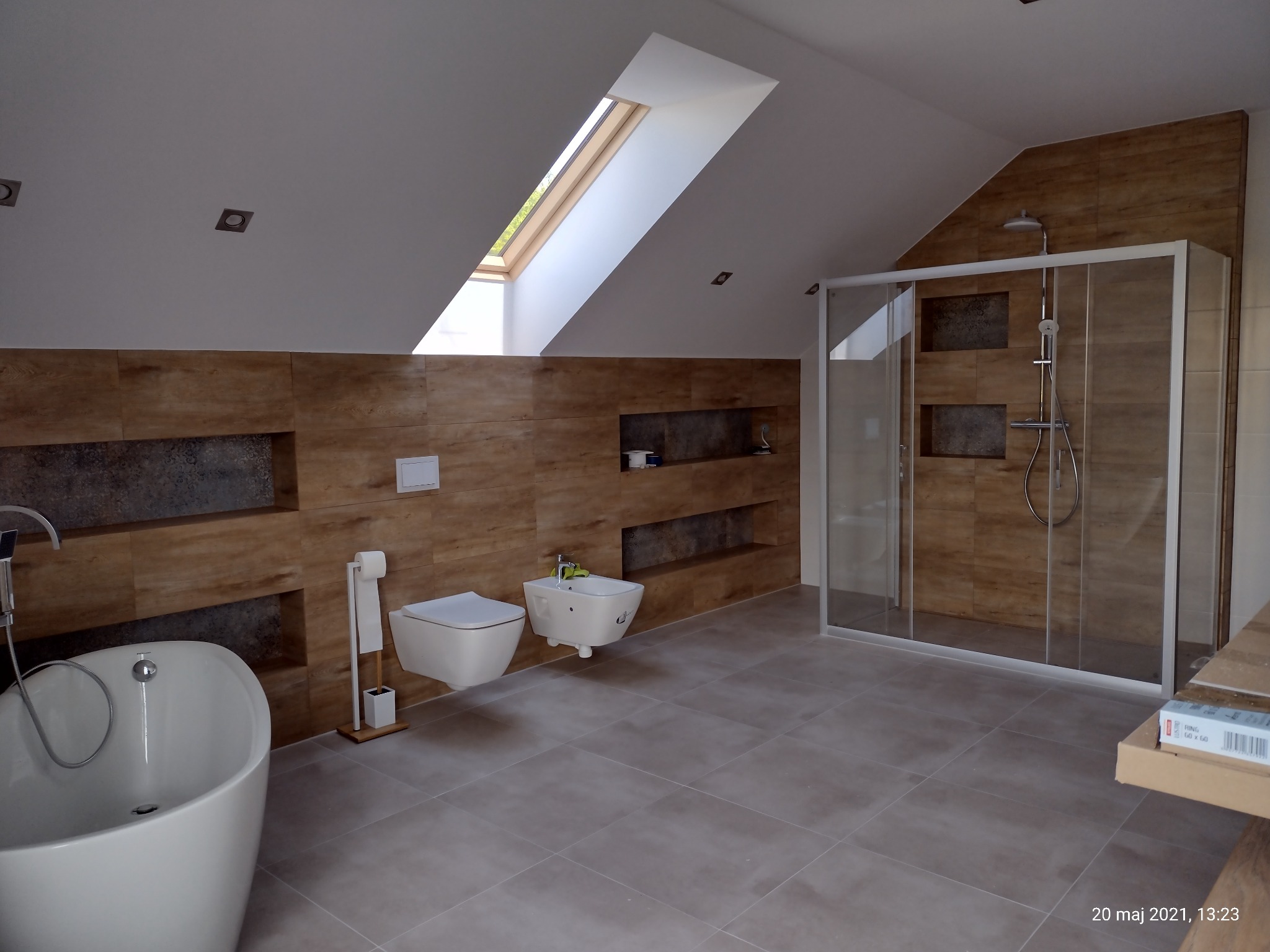 Nowoczesna łazienka na poddaszu z wanną wolnostojącą, toaletą, bidetem i kabiną prysznicową typu walk-in, wykończona płytkami w odcieniach brązu i szarości, oświetlona światłem dziennym z okna...