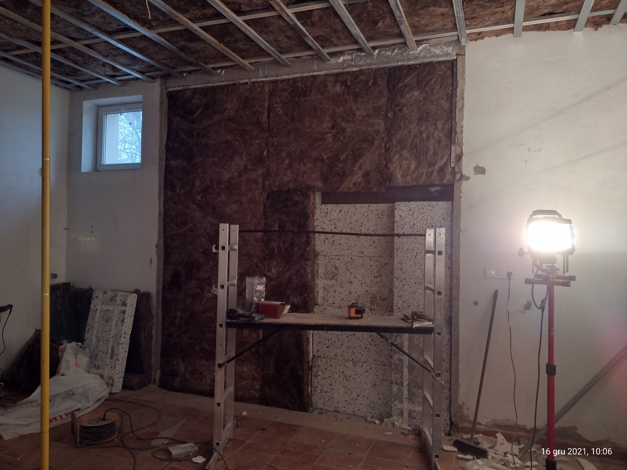 Wnętrze w trakcie prac remontowych: ściana z izolacją z wełny mineralnej, częściowo wykończona płyta, rusztowanie z narzędziami, lampa budowlana i widoczne elementy konstrukcji sufitu.