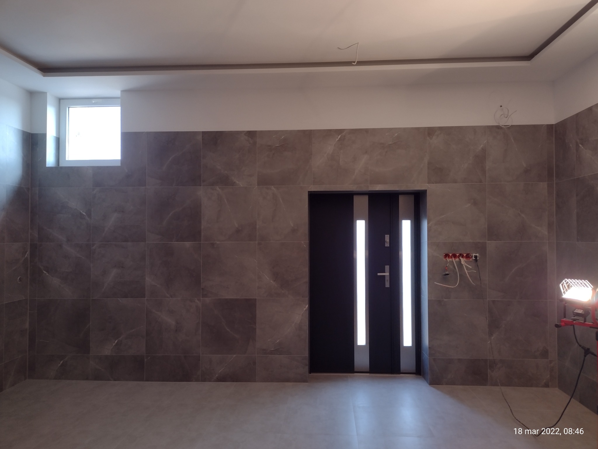 Wykończona łazienka z dużymi, szarymi płytkami imitującymi marmur na ścianach, podświetlona lampą roboczą. Widoczna nowa, czarna ościeżnica z drzwiami z dwoma pionowymi pasami szkła.