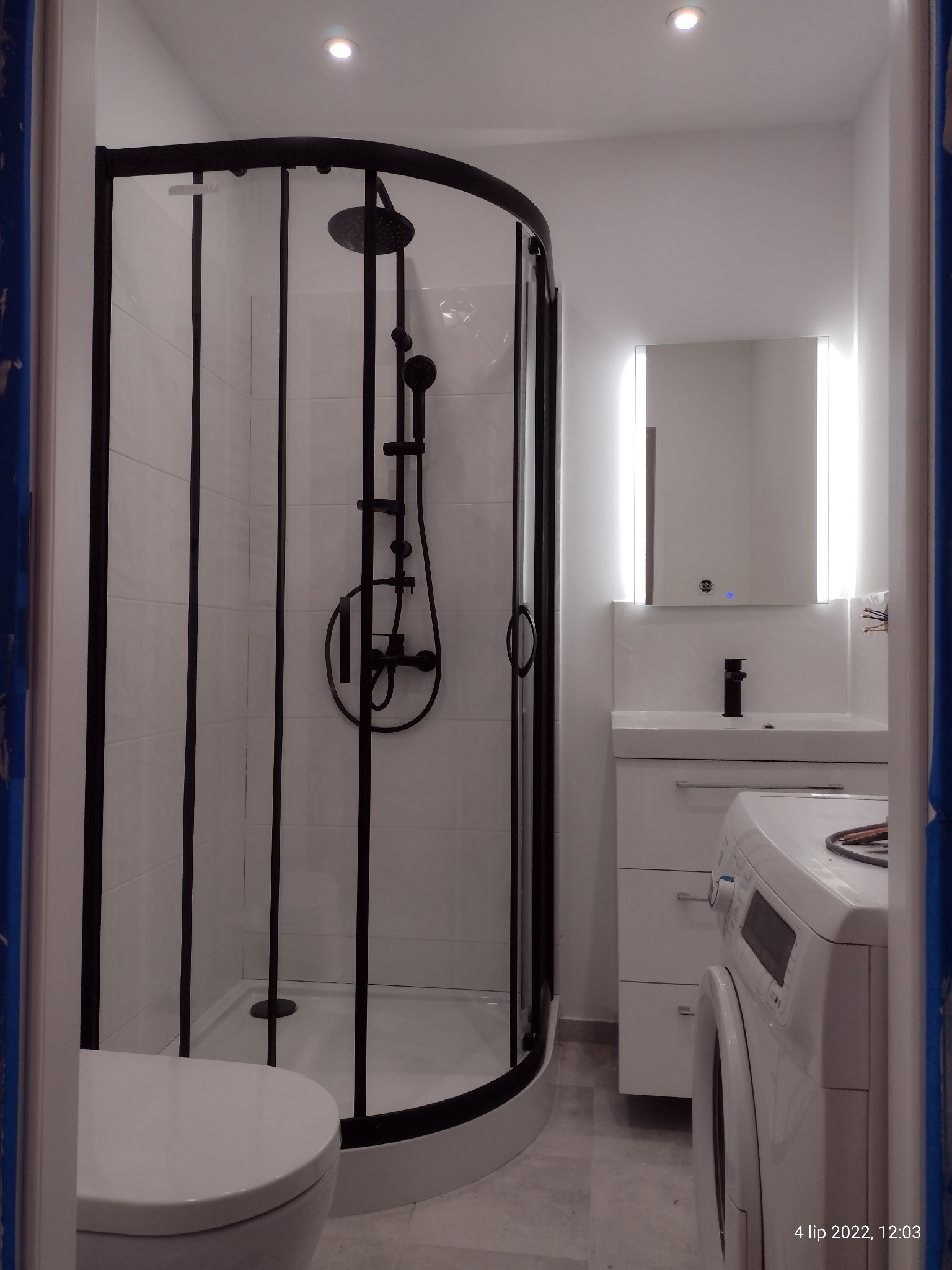 Wykończona łazienka z kabiną prysznicową w czarnej ramie, białą umywalką z szafką, lustrem z podświetleniem LED i pralką, widok zza toalety.