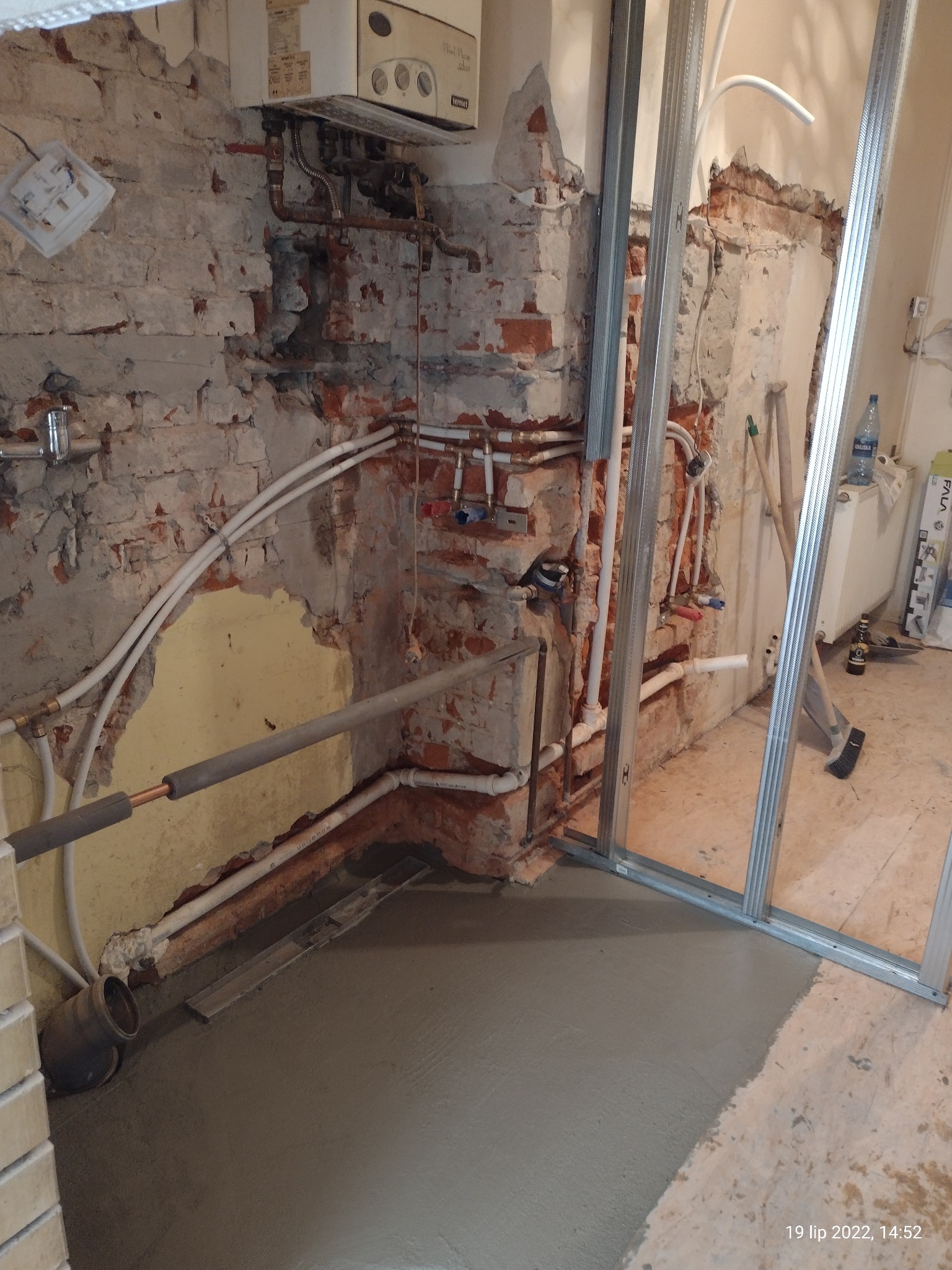 Surowa ściana z odsłoniętą cegłą, rurami i instalacją grzewczą, świeżo wylana wylewka, metalowe profile konstrukcyjne, etap prac wykończeniowych w pomieszczeniu.