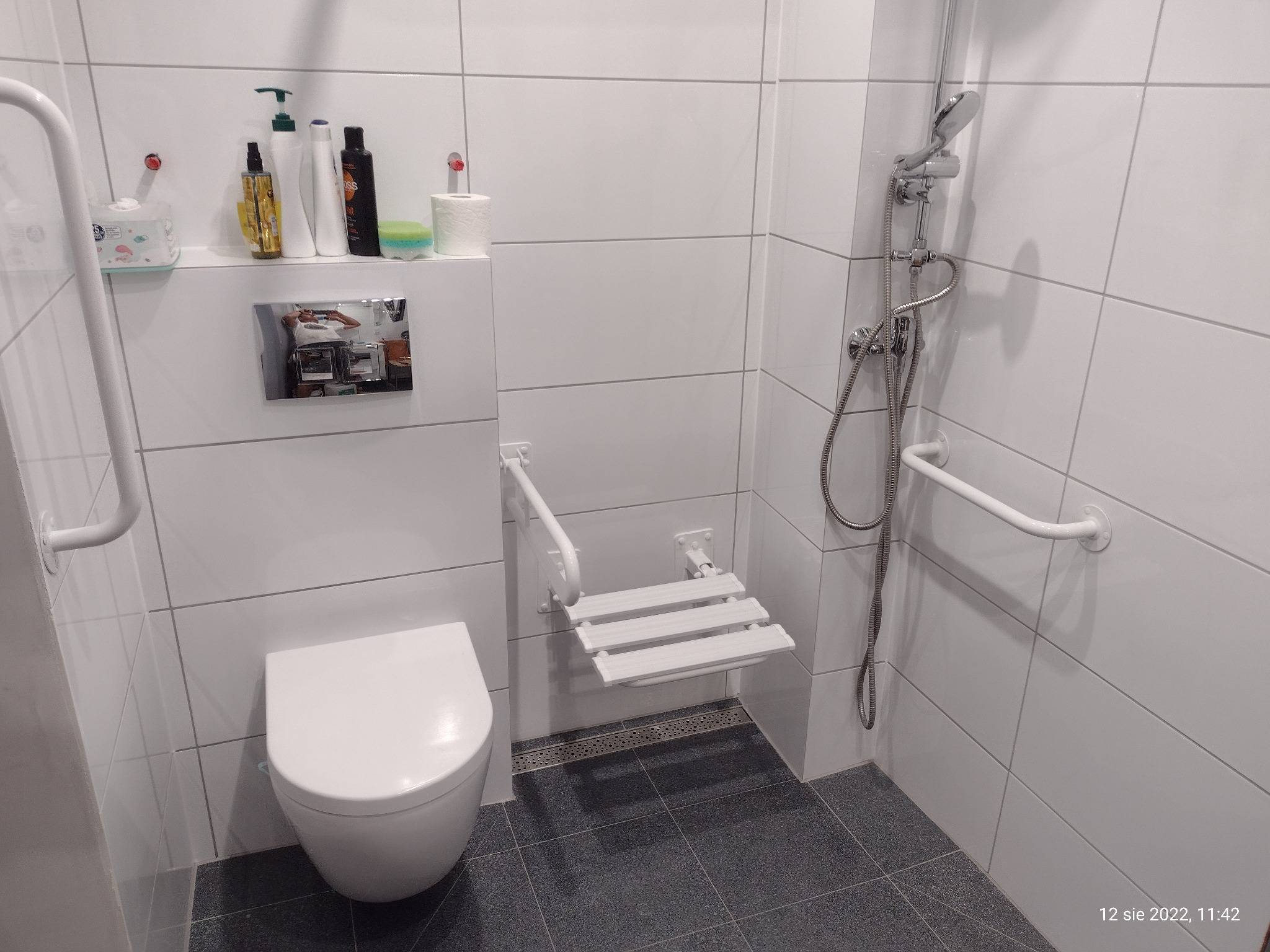 Biała łazienka z sedesem podwieszanym, składanym siedziskiem prysznicowym dla niepełnosprawnych i uchwytami, wykończona płytkami w odcieniach bieli i szarości.