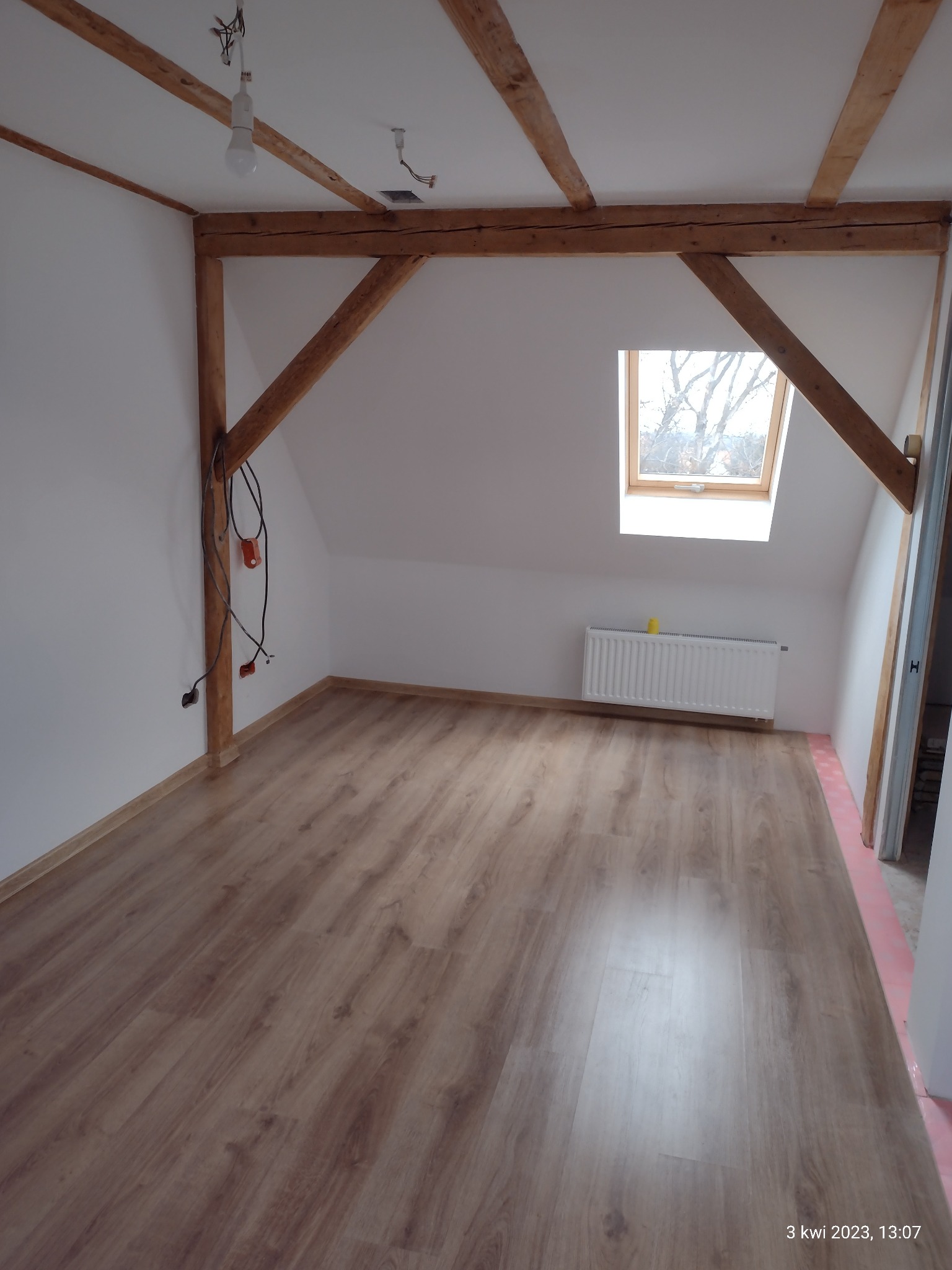 Poddasze z drewnianą konstrukcją stropu, nową podłogą laminowaną i oknem dachowym; widoczne kable elektryczne zwisające ze ściany, grzejnik pod oknem i przygotowane podłoże podłogowe przy ścianie.