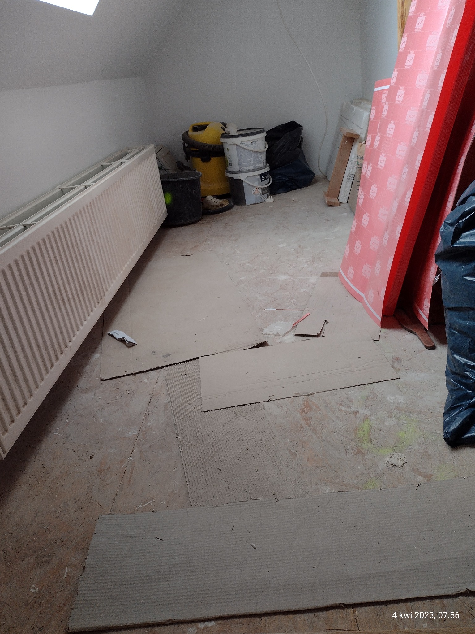 Przygotowania do prac wykończeniowych na poddaszu: zabezpieczona podłoga kartonem, grzejnik, wiadra z farbą, odkurzacz i płyty izolacyjne.