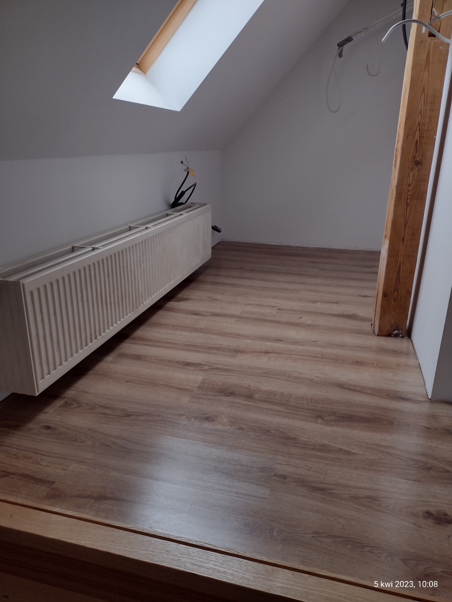 Poddasze w Świdnicy z drewnianą podłogą, grzejnikiem i oknem dachowym przed wykończeniem, widoczne kable i elementy konstrukcyjne.