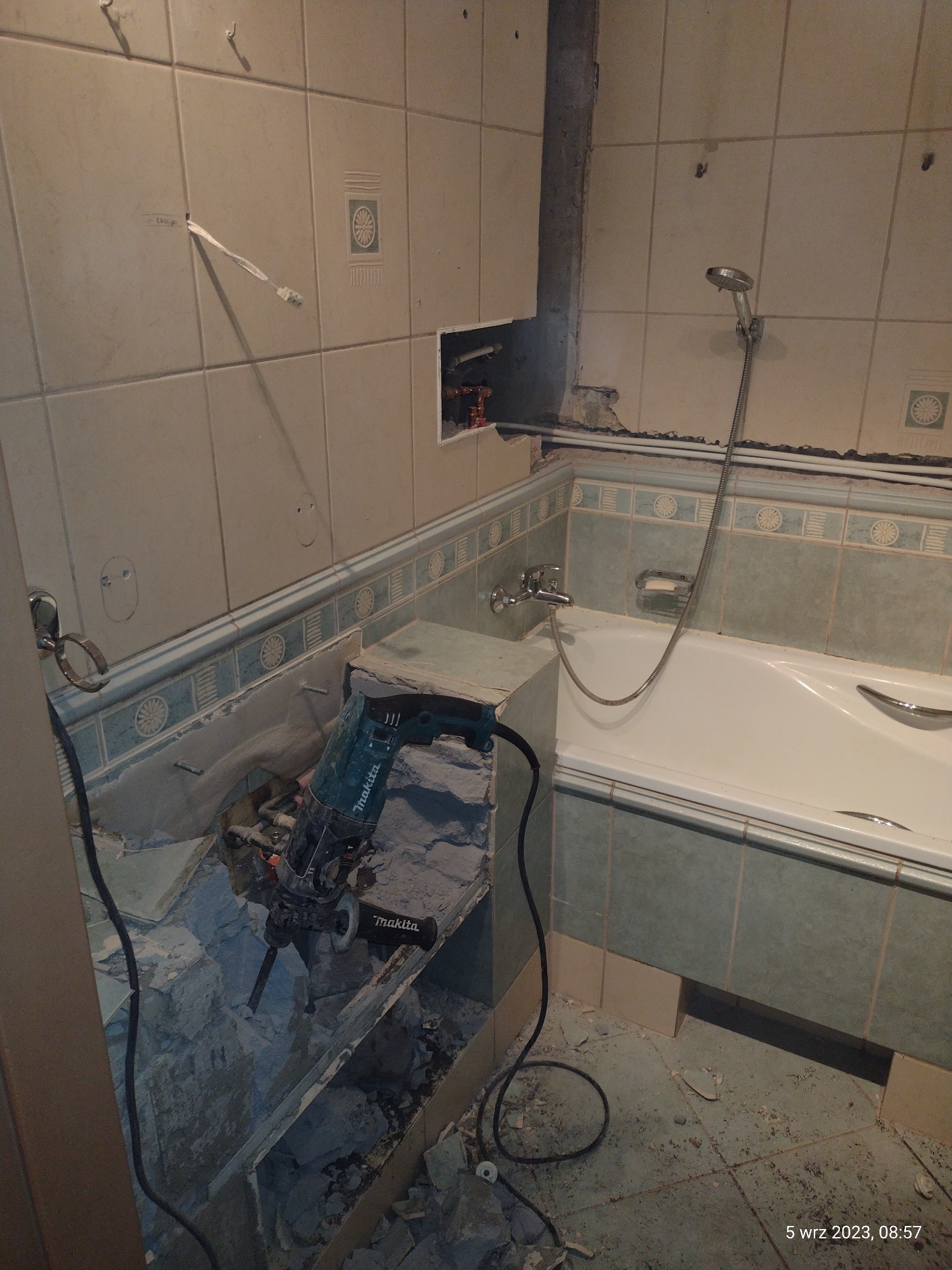 Demontaż starych płytek w łazience za pomocą młotowiertarki Makita, widoczne fragmenty instalacji hydraulicznej i elektrycznej, gruz na podłodze.