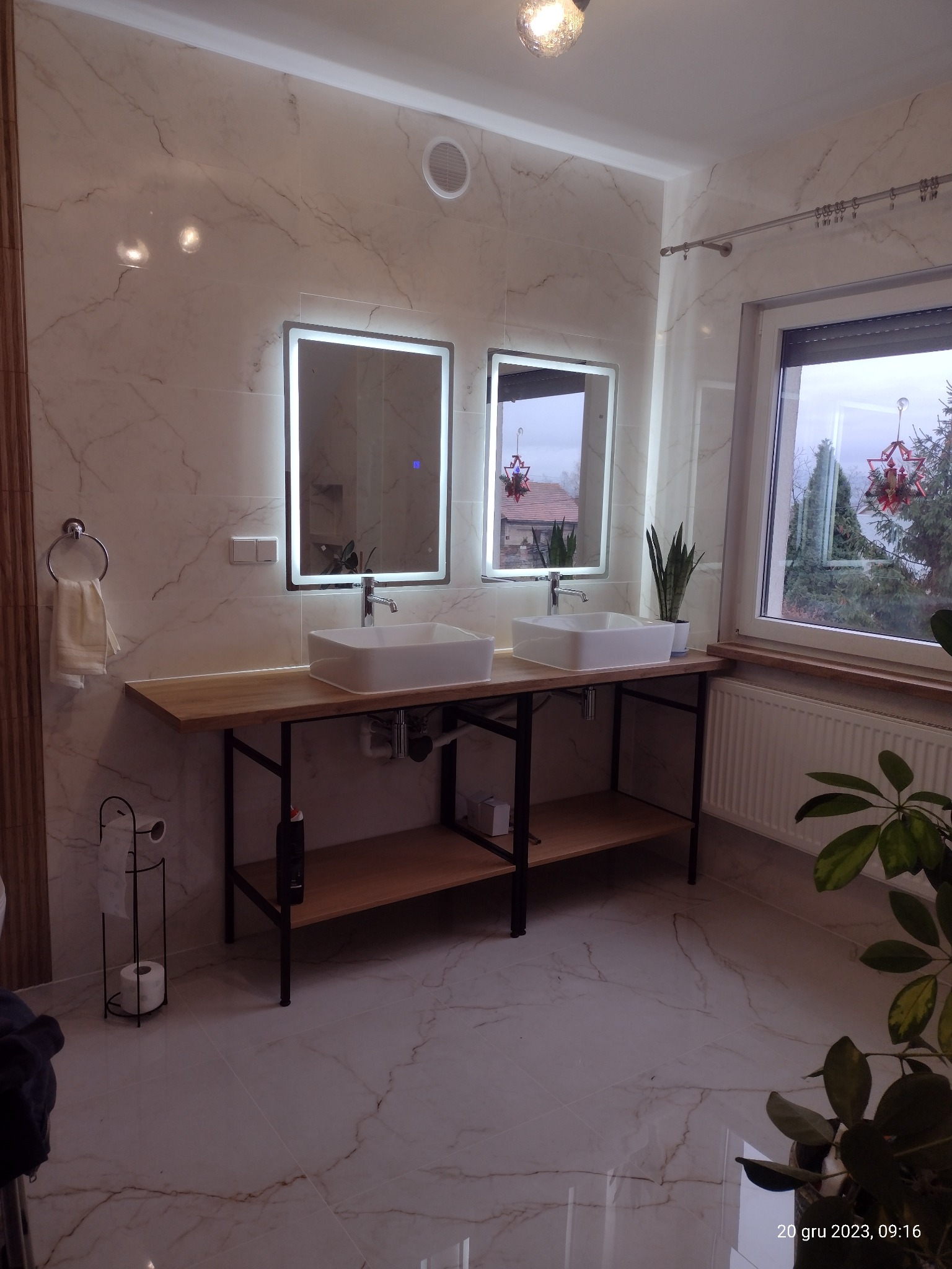 Nowoczesna łazienka z dwoma prostokątnymi umywalkami nablatowymi na drewnianym blacie, podświetlane lustra, marmurowe płytki na ścianach i podłodze, oraz dekoracje świąteczne widoczne za oknem.