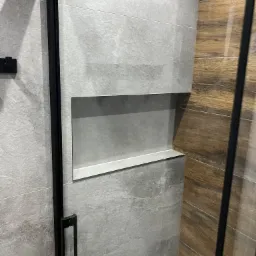 Fragment łazienki po remoncie: szare, kamienne płytki z wnęką, kontrastujące z drewnopodobnymi płytkami i czarną ramą kabiny prysznicowej.