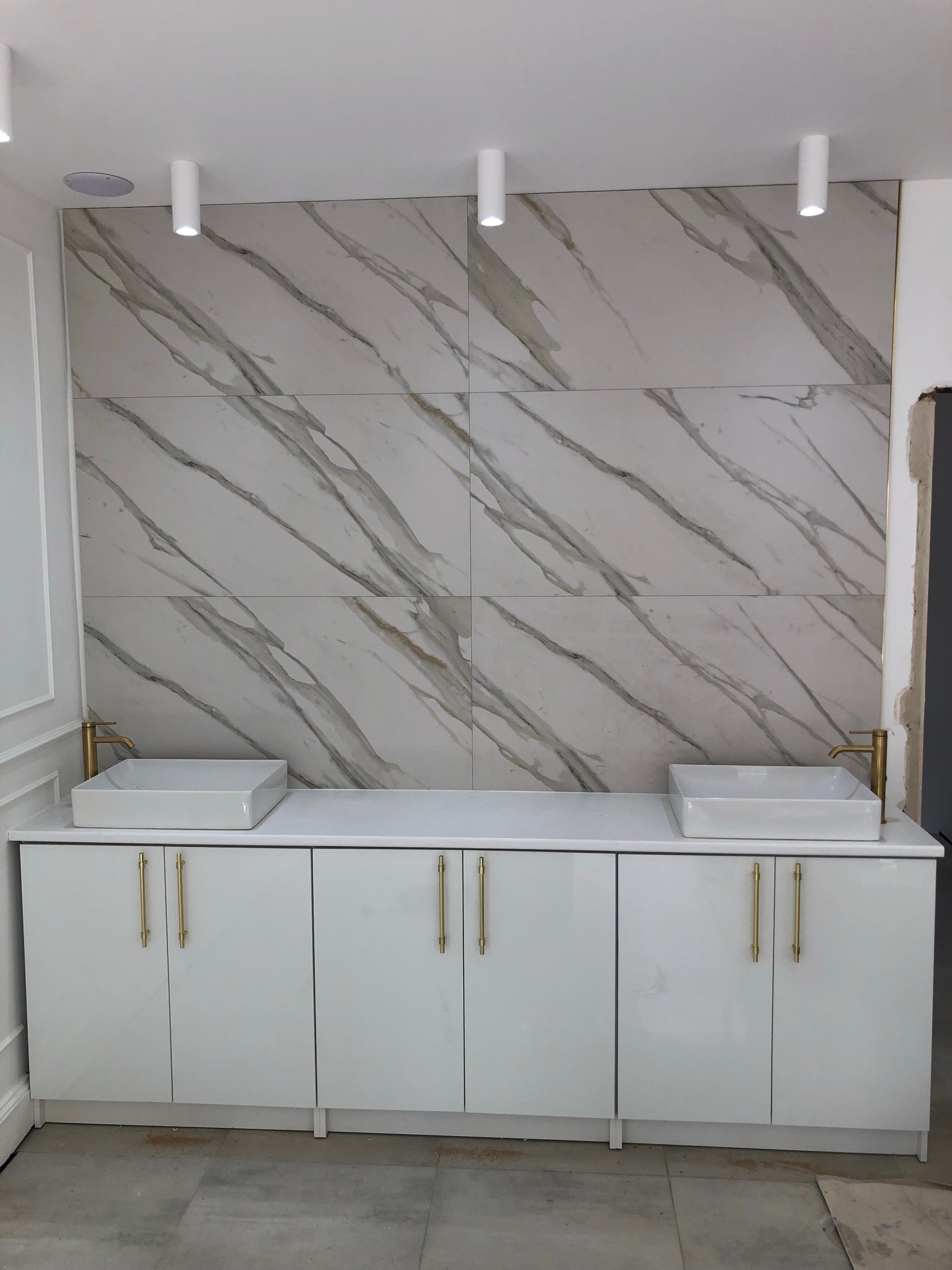 Nowoczesna łazienka w Starachowicach po remoncie: białe szafki pod umywalkami z prostokątnymi, nablatowymi umywalkami i złotymi bateriami, ściana wyłożona płytkami imitującymi marmur, oświetlenie...