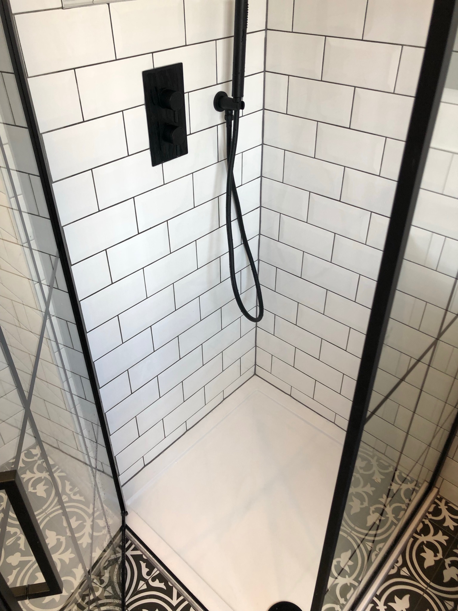 Kabina prysznicowa z czarnymi elementami armatury i białymi, prostokątnymi płytkami ułożonymi w cegiełkę, kontrastująca z wzorzystą, czarno-białą podłogą.