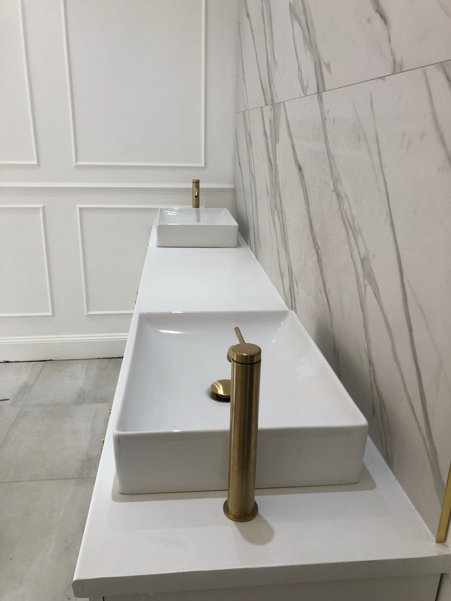 Dwie białe, prostokątne umywalki nablatowe z złotymi bateriami, ustawione na białym blacie. W tle białe ściany: jedna z dekoracyjnymi panelami, druga wykończona płytkami imitującymi marmur.