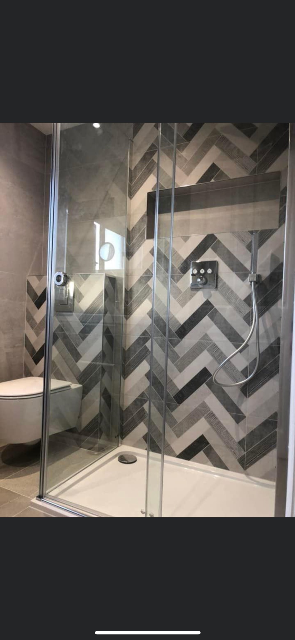 Nowoczesna kabina prysznicowa z deszczownicą i szarymi płytkami ułożonymi w jodełkę, z widoczną toaletą w tle i wnęką w ścianie.