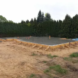 Wylana płyta fundamentowa z szalunkiem drewnianym i wystającymi prętami zbrojeniowymi na piaszczystym terenie, w tle rząd wysokich, gęstych tuj.