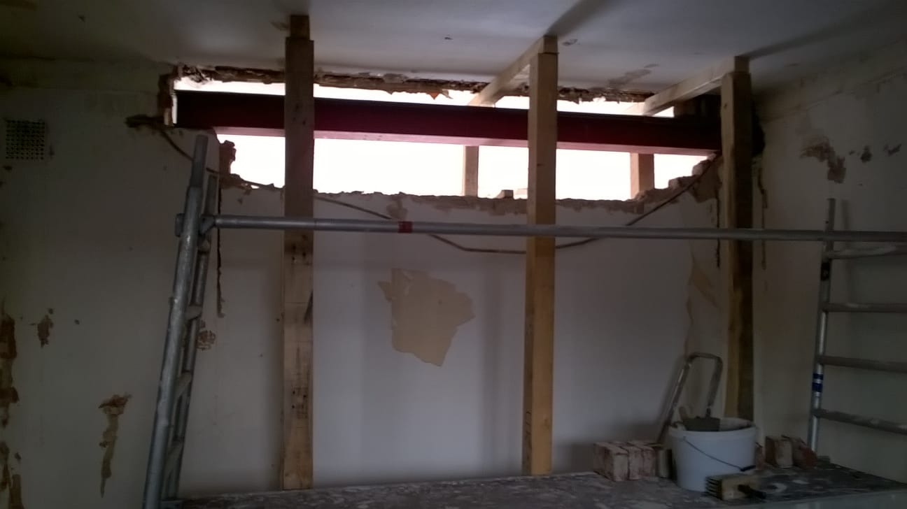 Wnętrze w trakcie prac remontowych z widoczną stalową belką podtrzymywaną przez drewniane stemple, fragmenty starego tynku na ścianach i sufitach, drabina i wiadro z zaprawą.