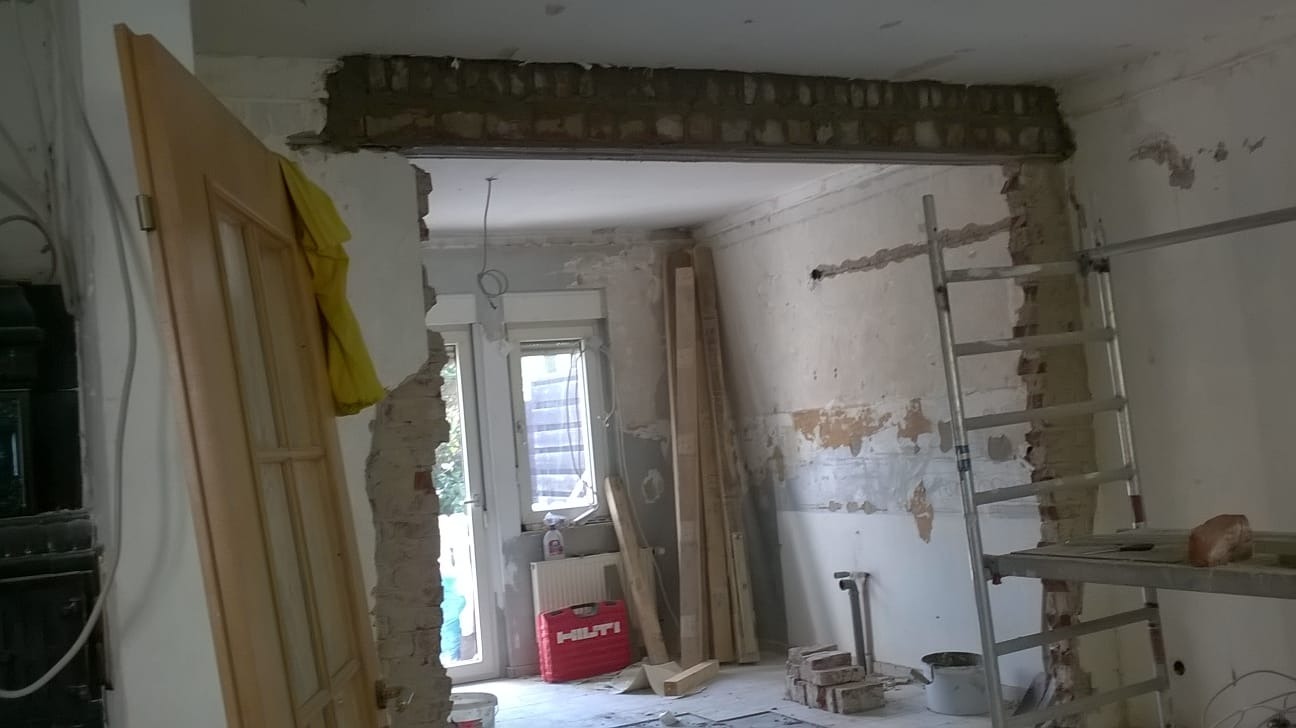Wnętrze w trakcie prac remontowych: odsłonięta cegła nad otworem drzwiowym, ściany z widocznymi warstwami farby, drabina, drzwi oparte o ścianę, narzędzia i materiały budowlane.