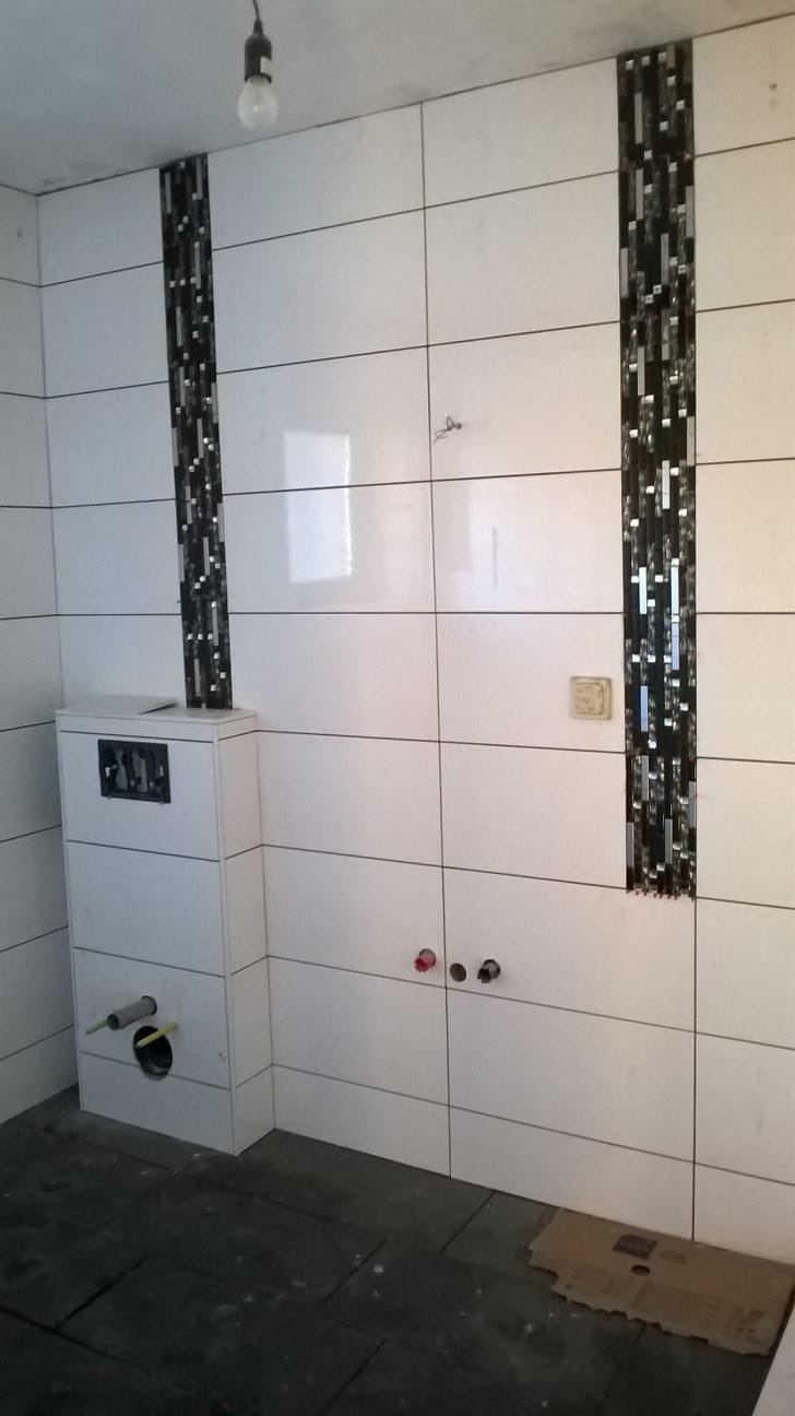 Wnętrze łazienki w trakcie remontu: białe, prostokątne płytki z dekoracyjnymi pasami czarnej mozaiki, zabudowa WC z widocznymi rurami, surowa podłoga.