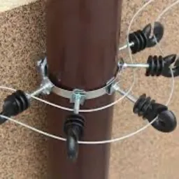 Brązowa rura spustowa z zamocowanym metalowym uchwytem i sześcioma czarnymi odgromnikami, zabezpieczonymi białymi linkami, na tle beżowej ściany z fakturą.