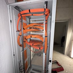 Gold Time Mykola Naboka - Pomarańczowe kable elektryczne uporządkowane w szafie rack, zabezpieczone czarnymi opaskami zaciskowymi, widoczne elementy konstrukcyjne szafy i fragment surowego wnętrza budynku w tle.