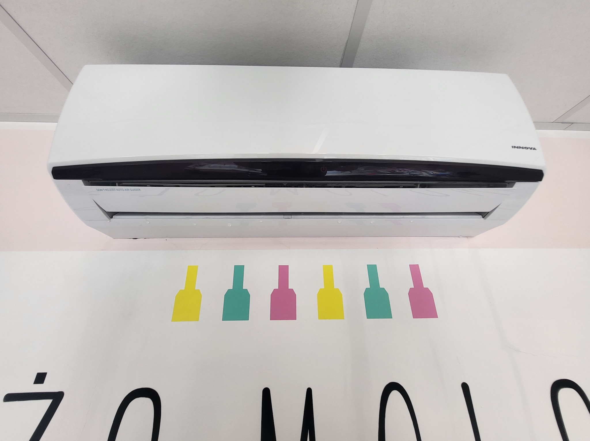 Biały klimatyzator marki Innova zamontowany na ścianie z kolorowymi grafikami w kształcie butelek