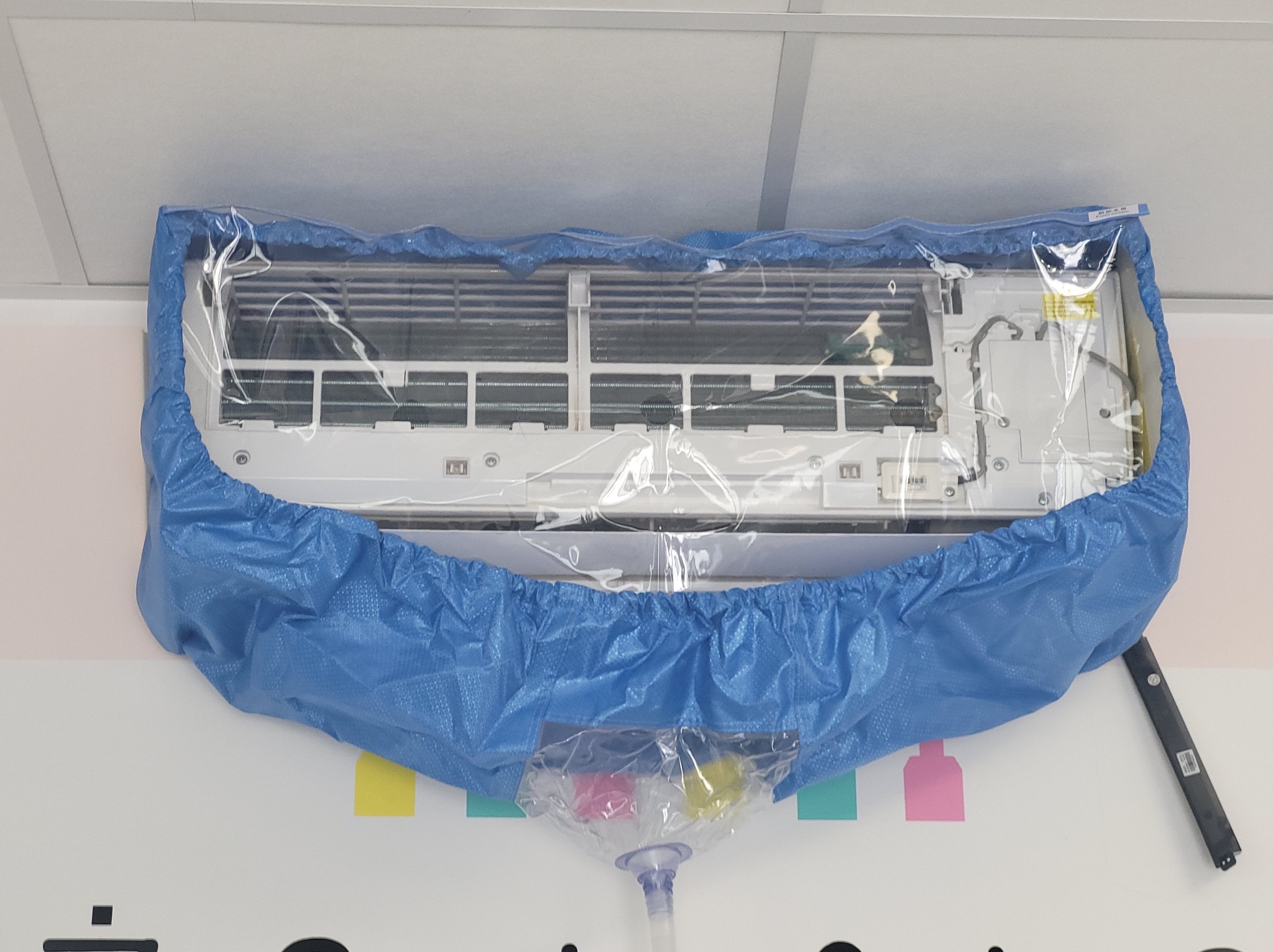 Klimatyzator ścienny zabezpieczony niebieską folią ochronną podczas czyszczenia, widoczny zbiornik na zanieczyszczenia z kolorowymi osadami.