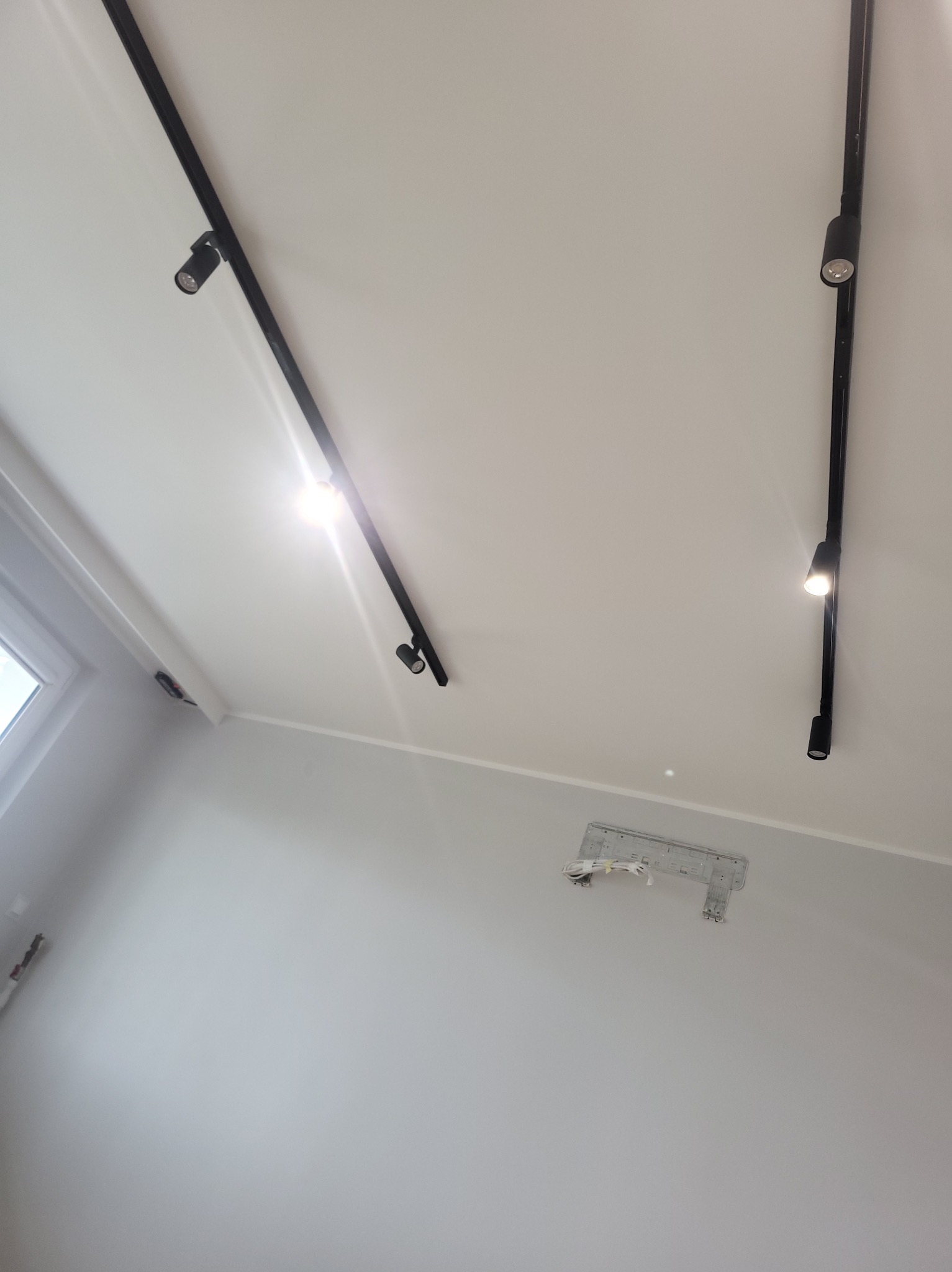 Biały sufit z zamontowanymi czarnymi szynami oświetleniowymi z reflektorami punktowymi oraz wspornik montażowy klimatyzatora na ścianie, widok z dołu.