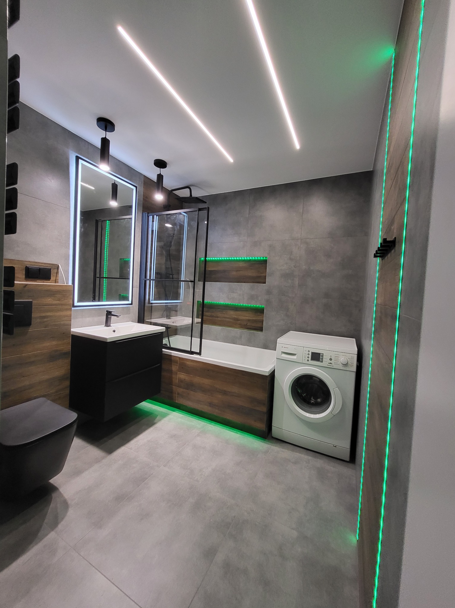 Nowoczesna łazienka z szarymi płytkami, drewnopodobnymi akcentami, podświetleniem LED, czarną armaturą i pralką Bosch.