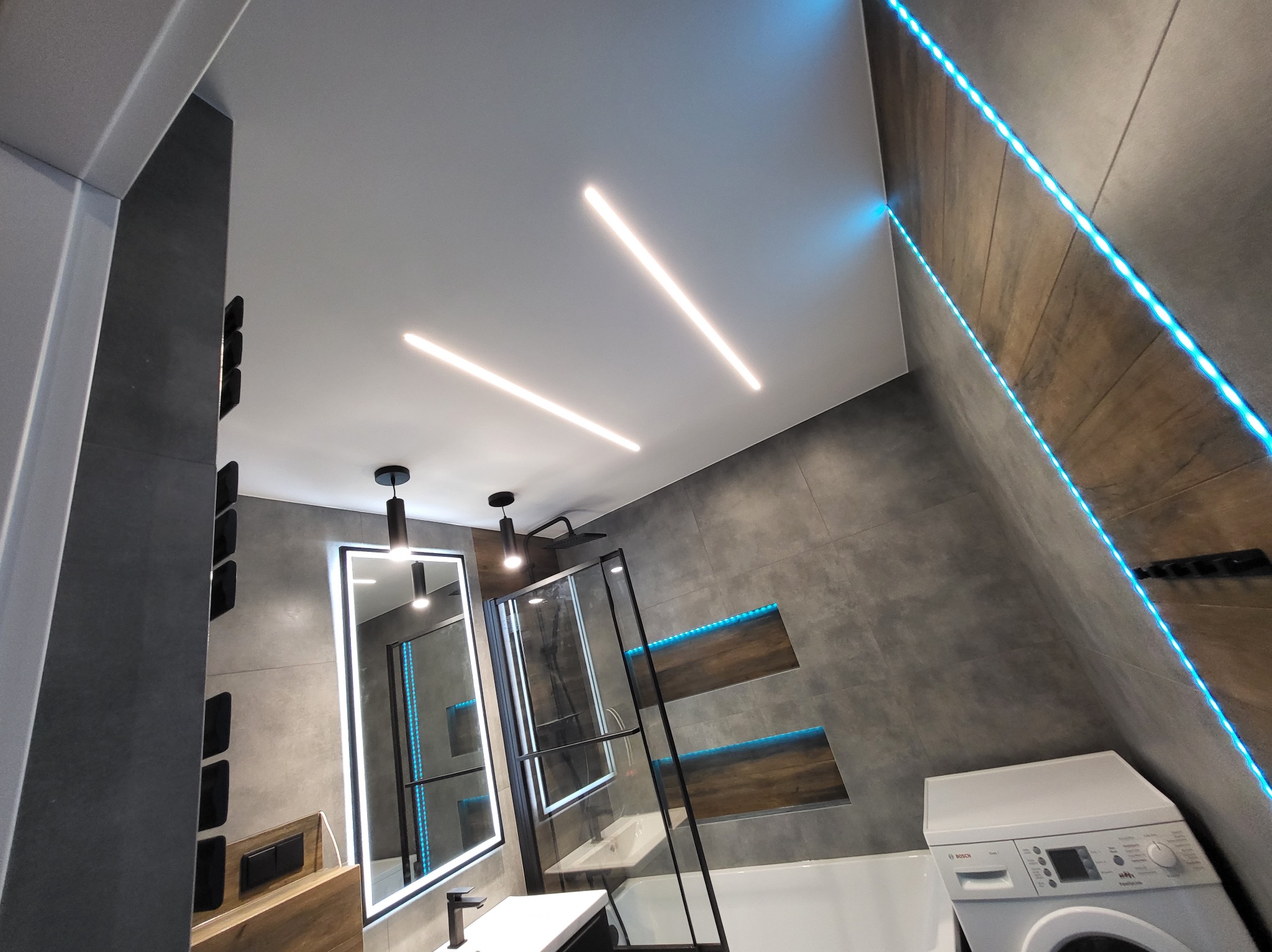 Nowoczesna łazienka z podświetleniem LED: liniowe lampy na suficie, niebieskie paski LED w niszach ściennych, lustro z podświetleniem i kabina prysznicowa ze szkła hartowanego.