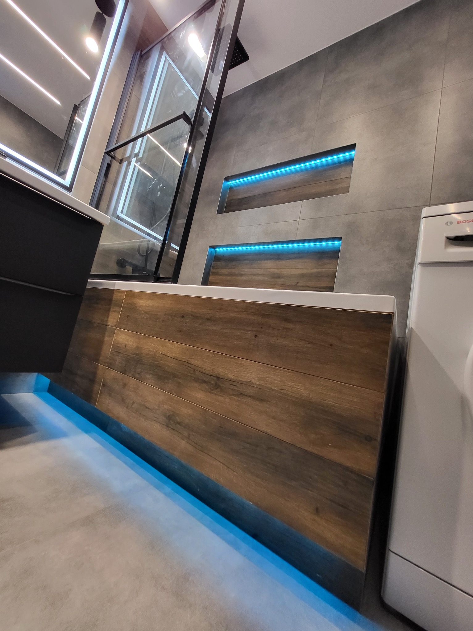 Nowoczesna łazienka z podświetlaną wanną obłożoną drewnopodobnymi panelami, kabina prysznicowa z czarnymi profilami, szare płytki ścienne z niszami podświetlonymi niebieskim światłem LED, pralka...