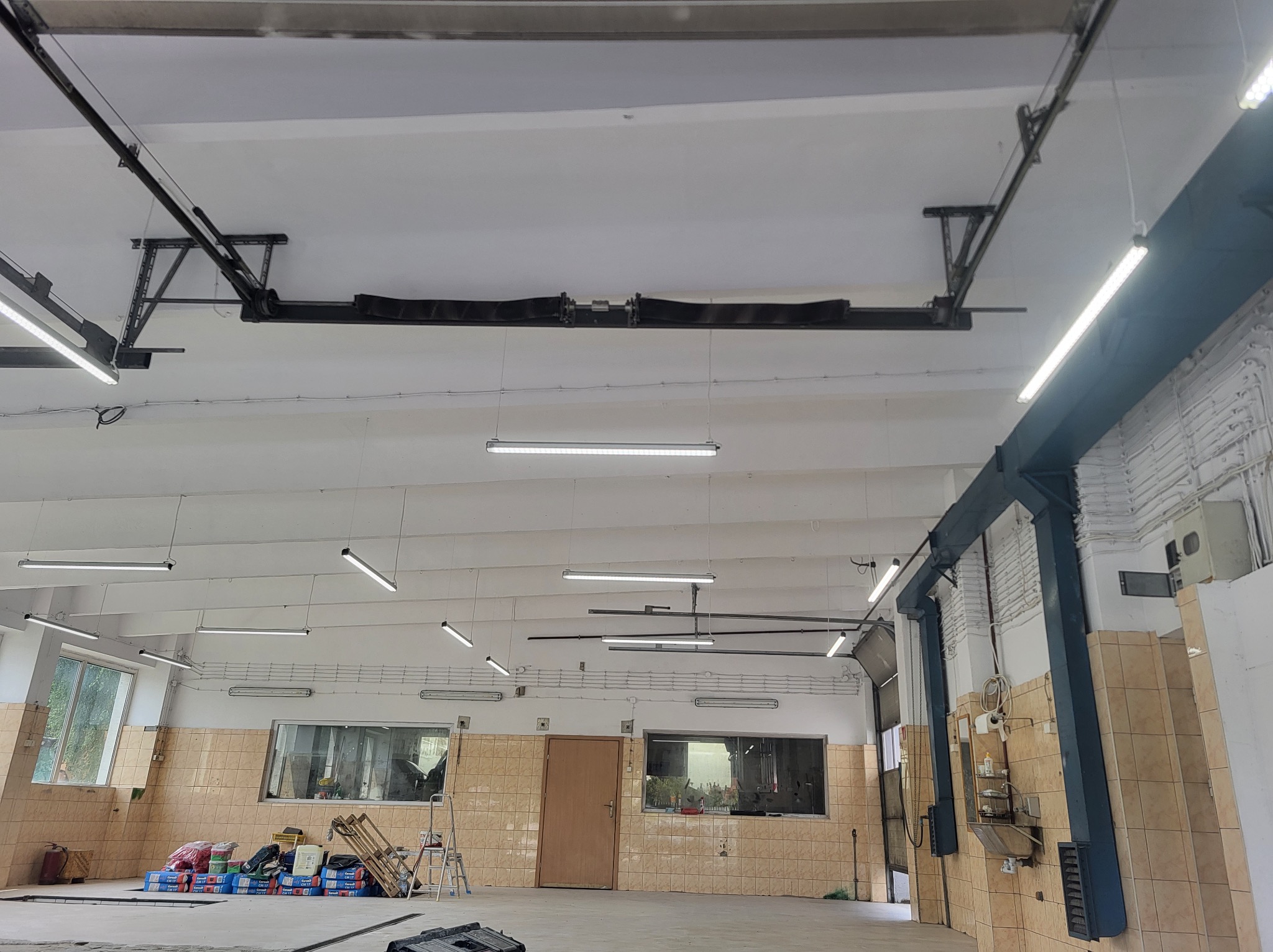 Wnętrze warsztatu z oświetleniem LED, widoczne podnośniki samochodowe, instalacje elektryczne na suficie i okna z widokiem na zewnątrz.